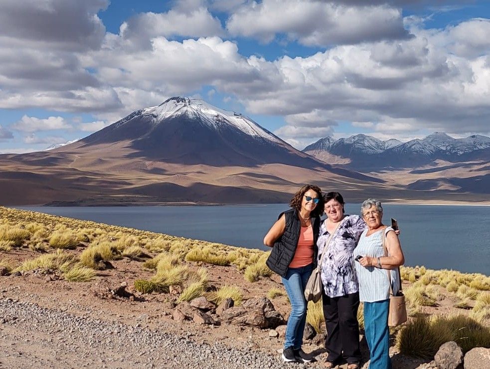 Tres personas posando junto a un lago con una montaña nevada y un cielo nublado al fondo.