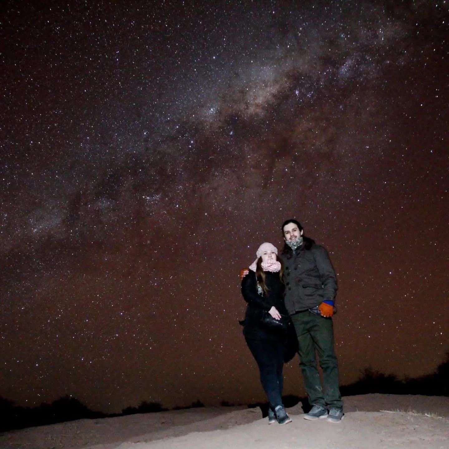 Dos personas de pie juntas bajo un cielo nocturno estrellado en un paisaje nevado.