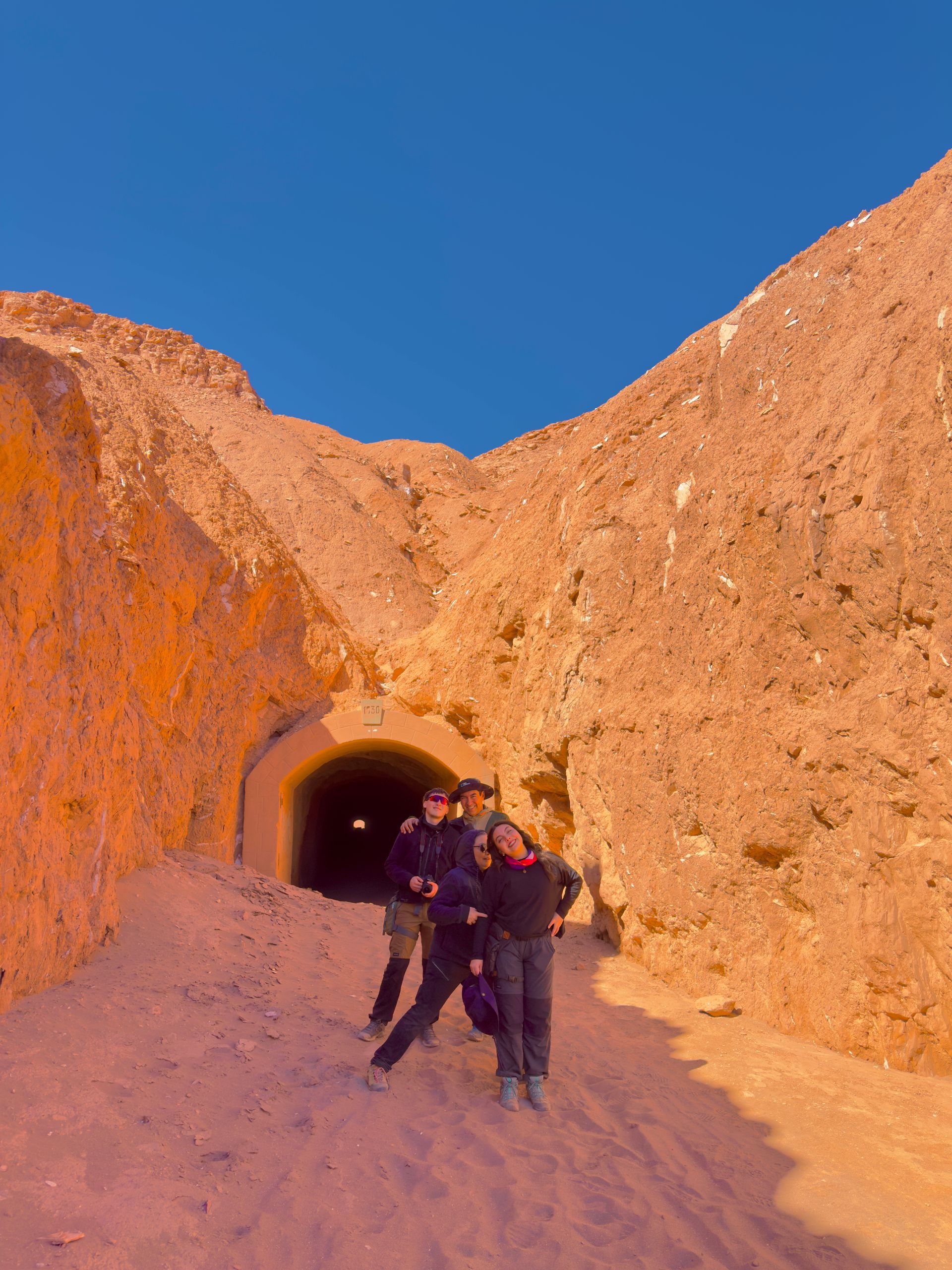 Dos personas de pie en la entrada de una cueva en un paisaje desértico rocoso iluminado por el sol.