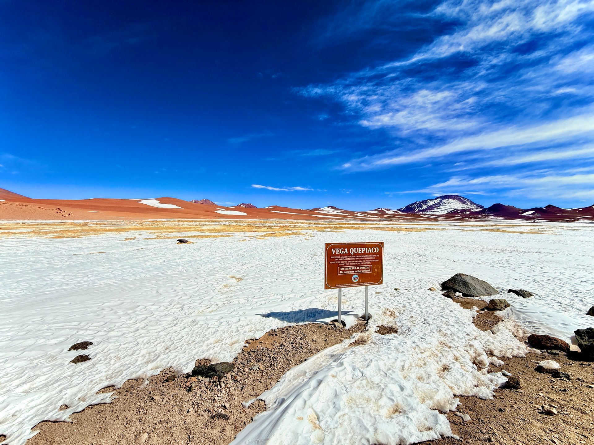 Paisaje árido cubierto de nieve con un pequeño letrero, montañas a lo lejos y un cielo azul intenso.