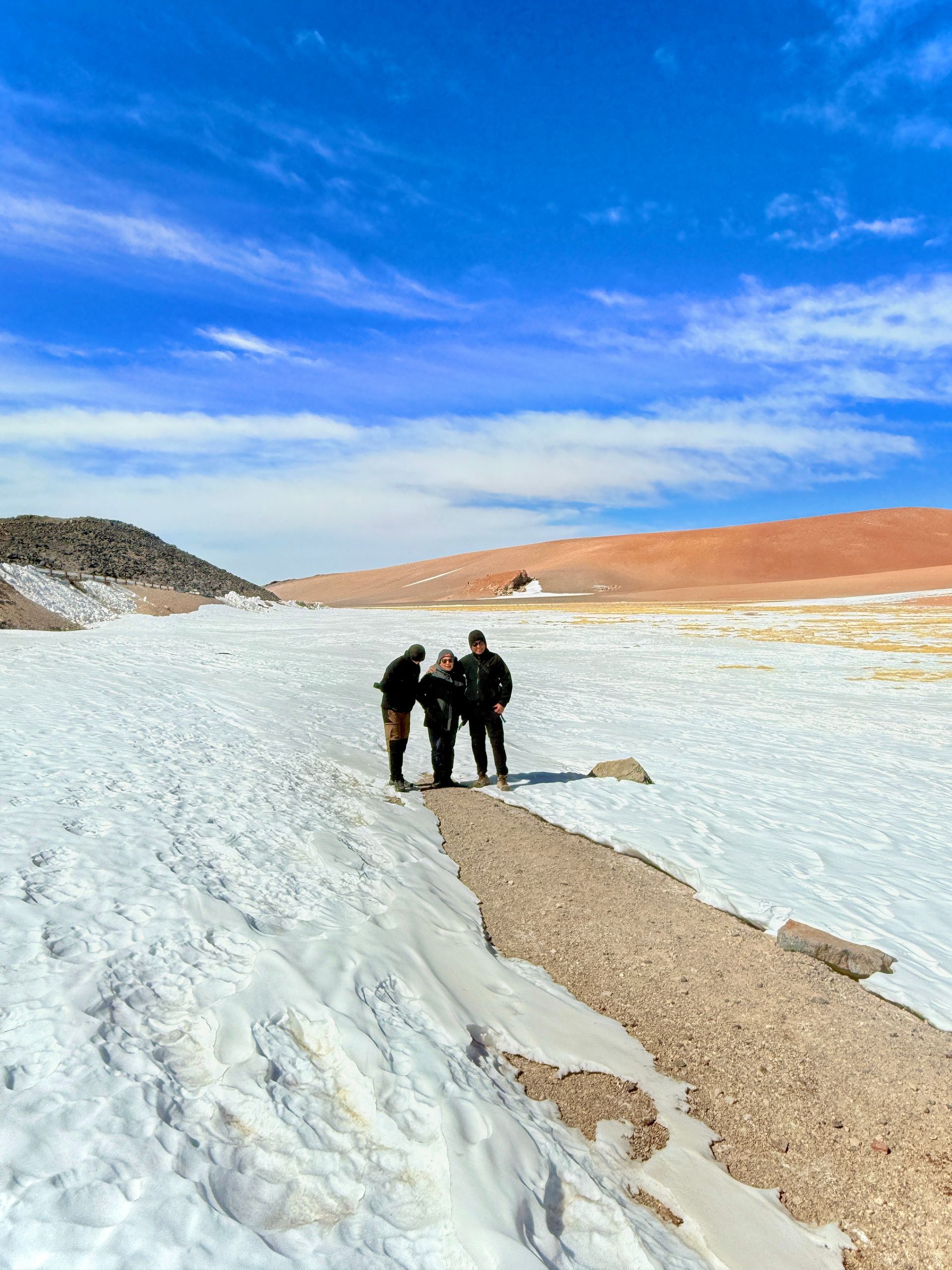 Tres personas caminan sobre una llanura salina blanca bajo un cielo azul, con colinas anaranjadas en la distancia.