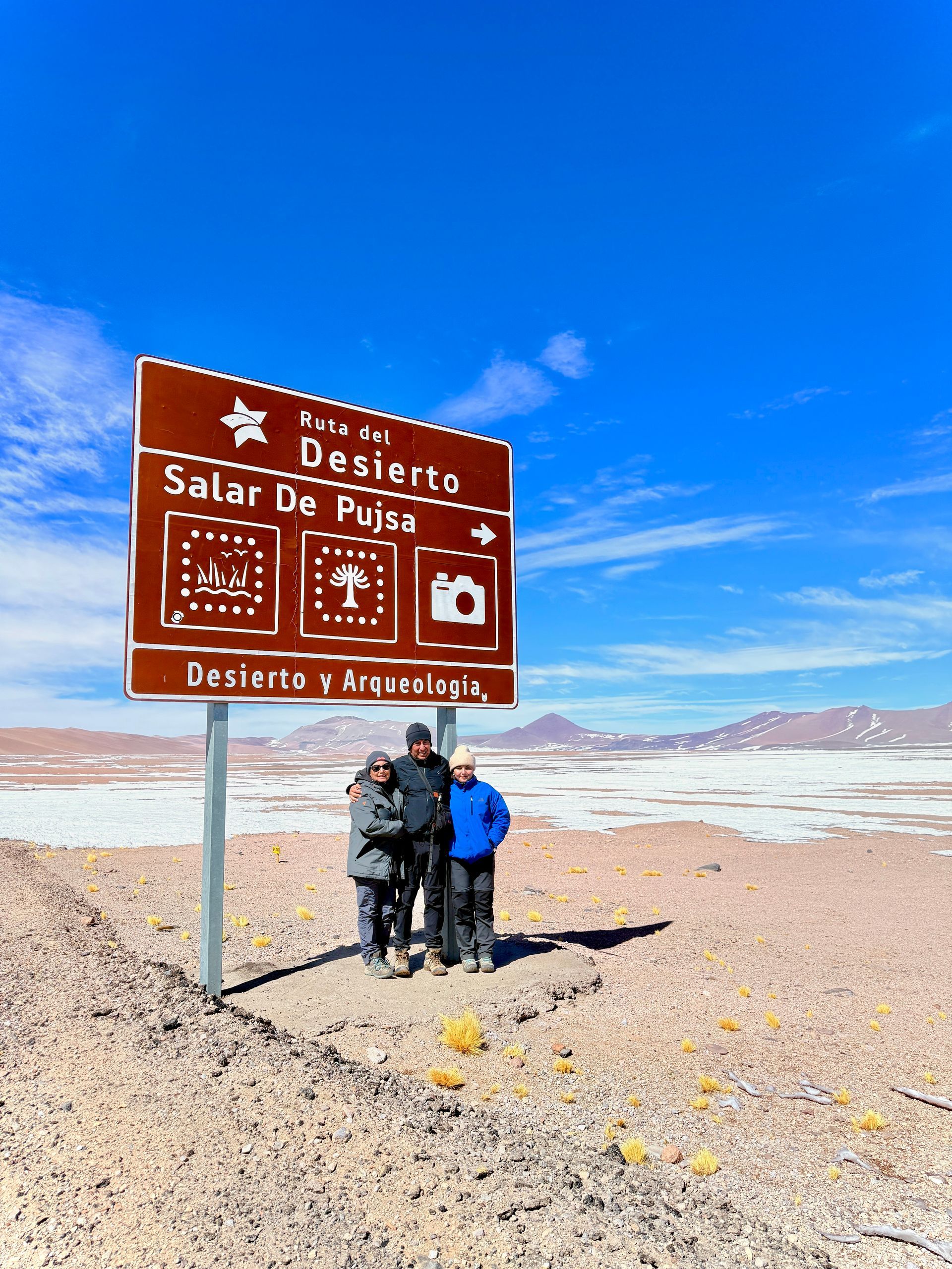Dos excursionistas junto a un cartel marrón del desierto de Salar de Uyuni bajo un cielo azul brillante.