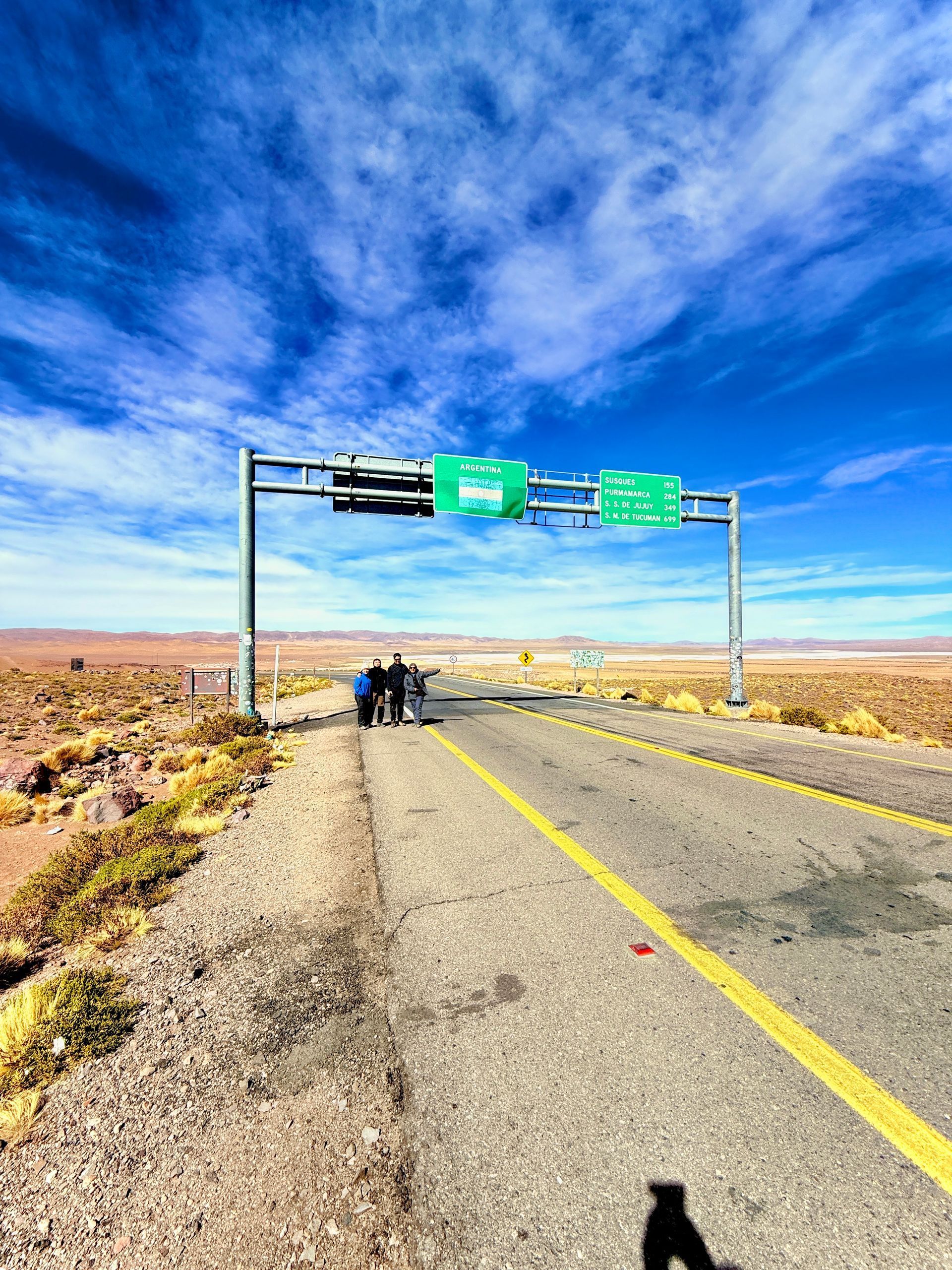 Carretera desértica bajo un cielo azul con señales aéreas y algunas personas de pie en la carretera.
