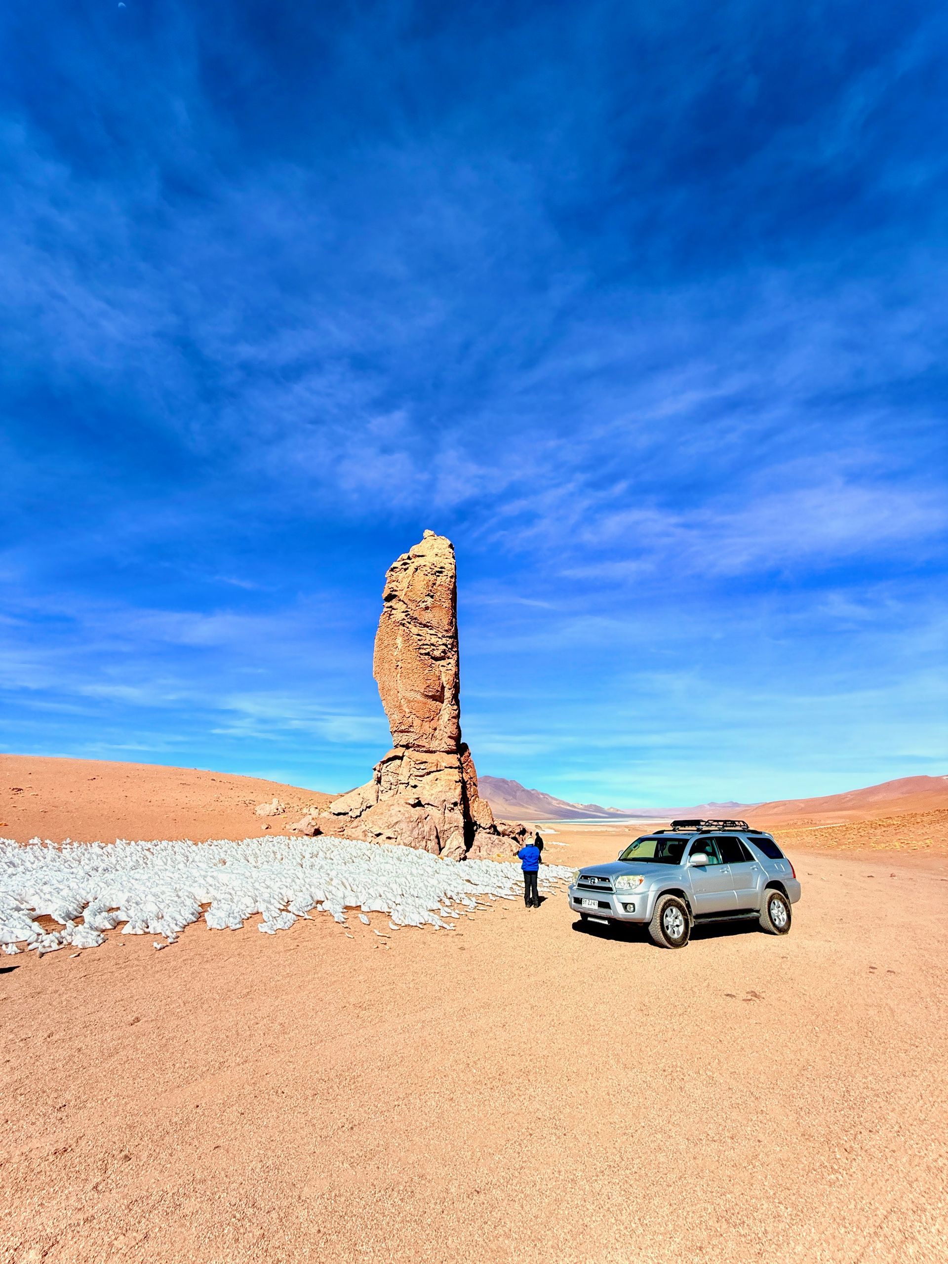 Un todoterreno cerca de una alta aguja rocosa en un desierto bajo un cielo azul intenso.