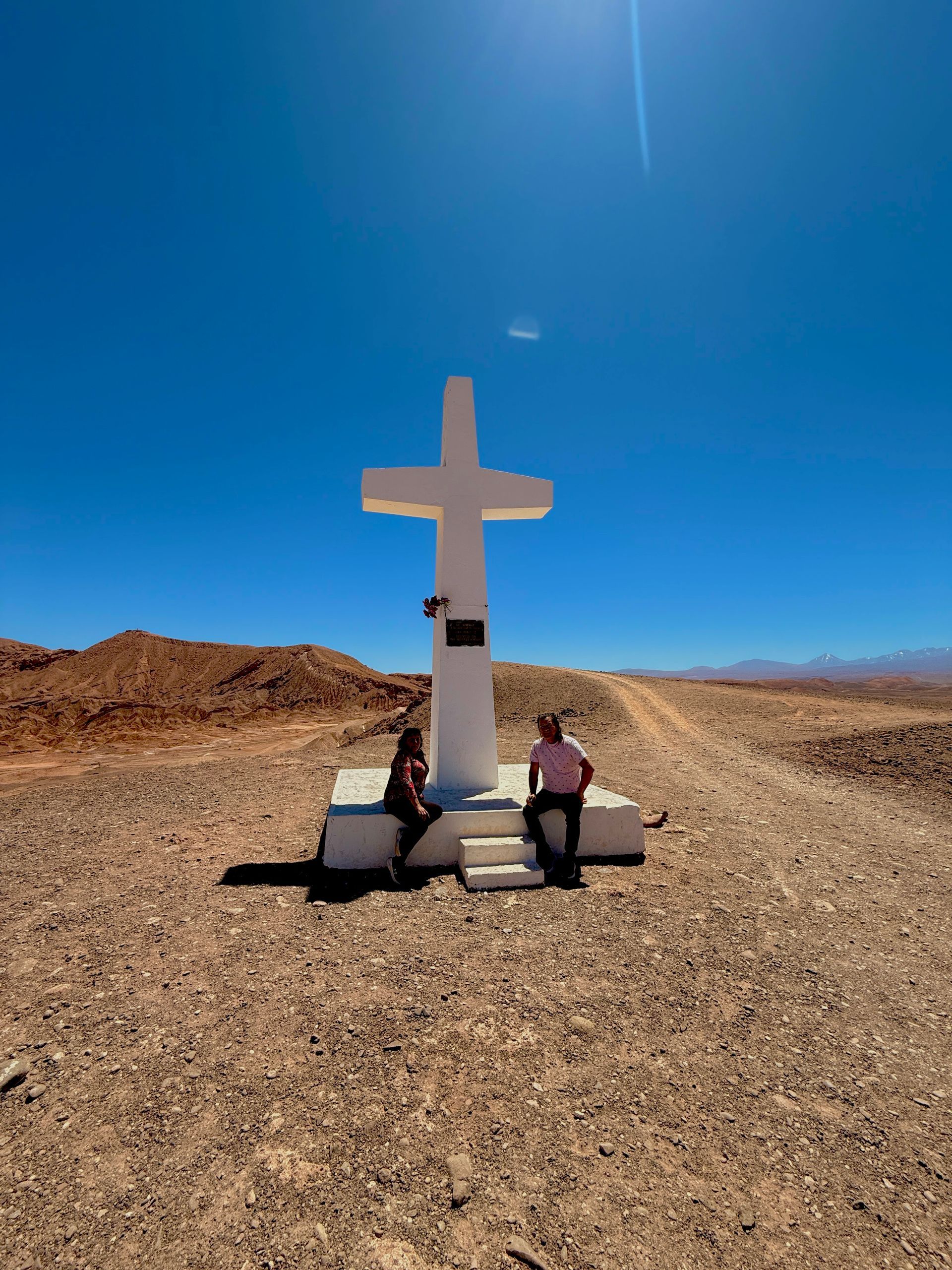Dos personas sentadas junto a un gran monumento con forma de cruz blanca en un desierto árido bajo un cielo azul despejado.