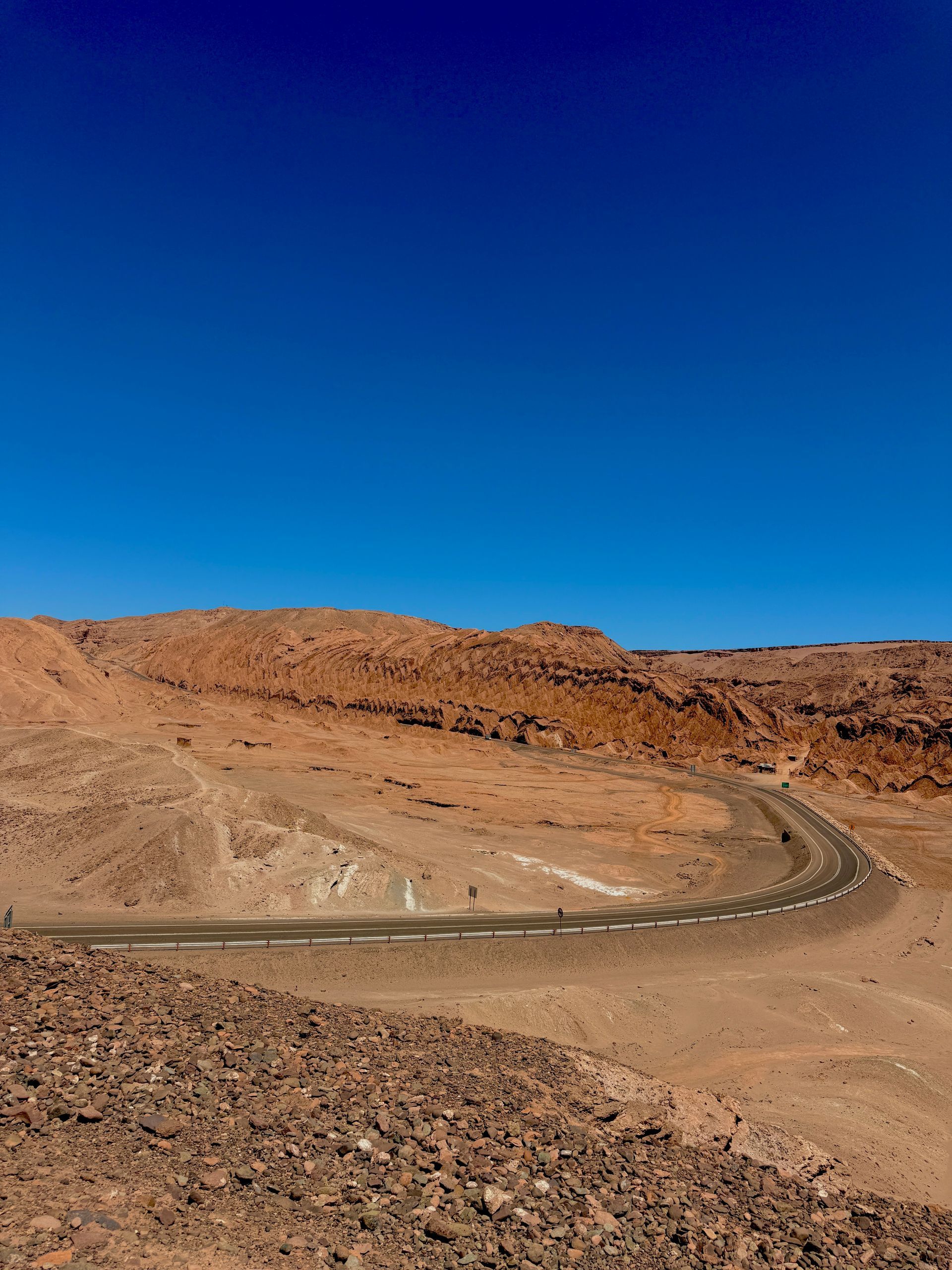 Carretera desértica serpenteando entre colinas rocosas de color marrón claro bajo un cielo azul profundo y despejado.