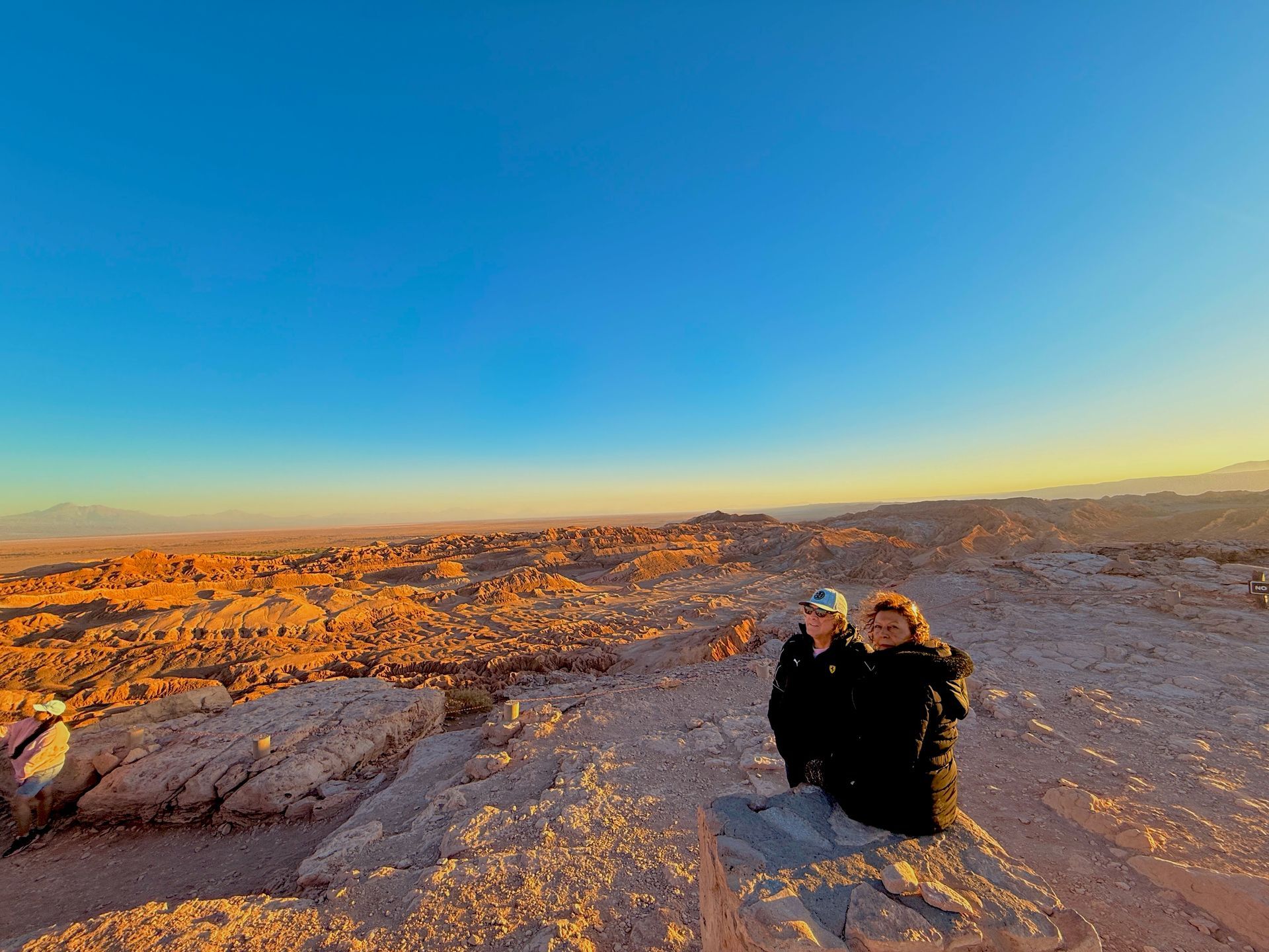 Dos excursionistas sentados en una meseta rocosa del desierto bajo un cielo azul despejado al atardecer.