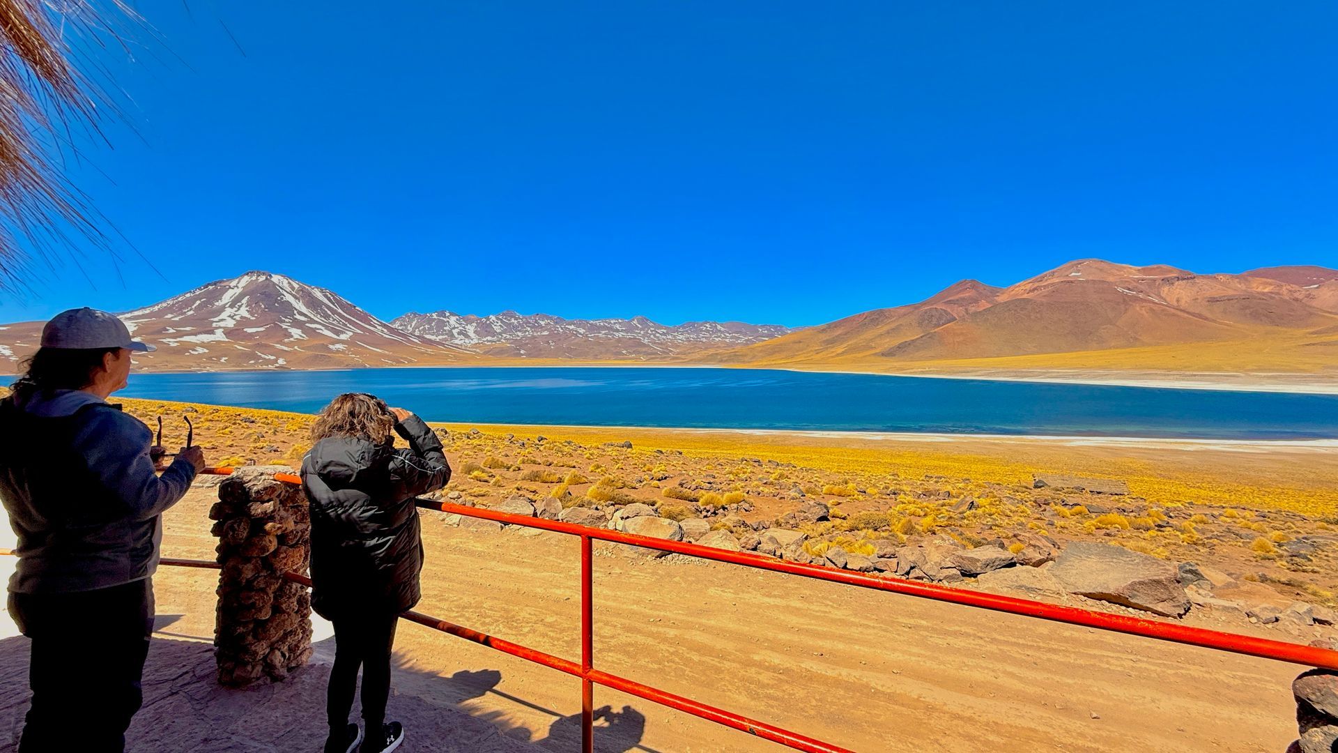 Gente contemplando un lago azul y colinas naranjas desde un mirador con barandilla roja bajo un cielo despejado.