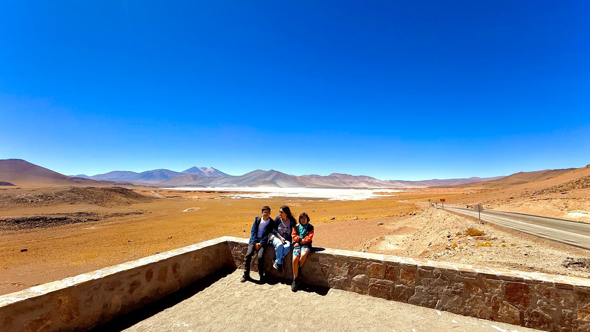 Tres personas de pie junto a un muro al borde de la carretera en un vasto desierto bajo un cielo azul brillante.
