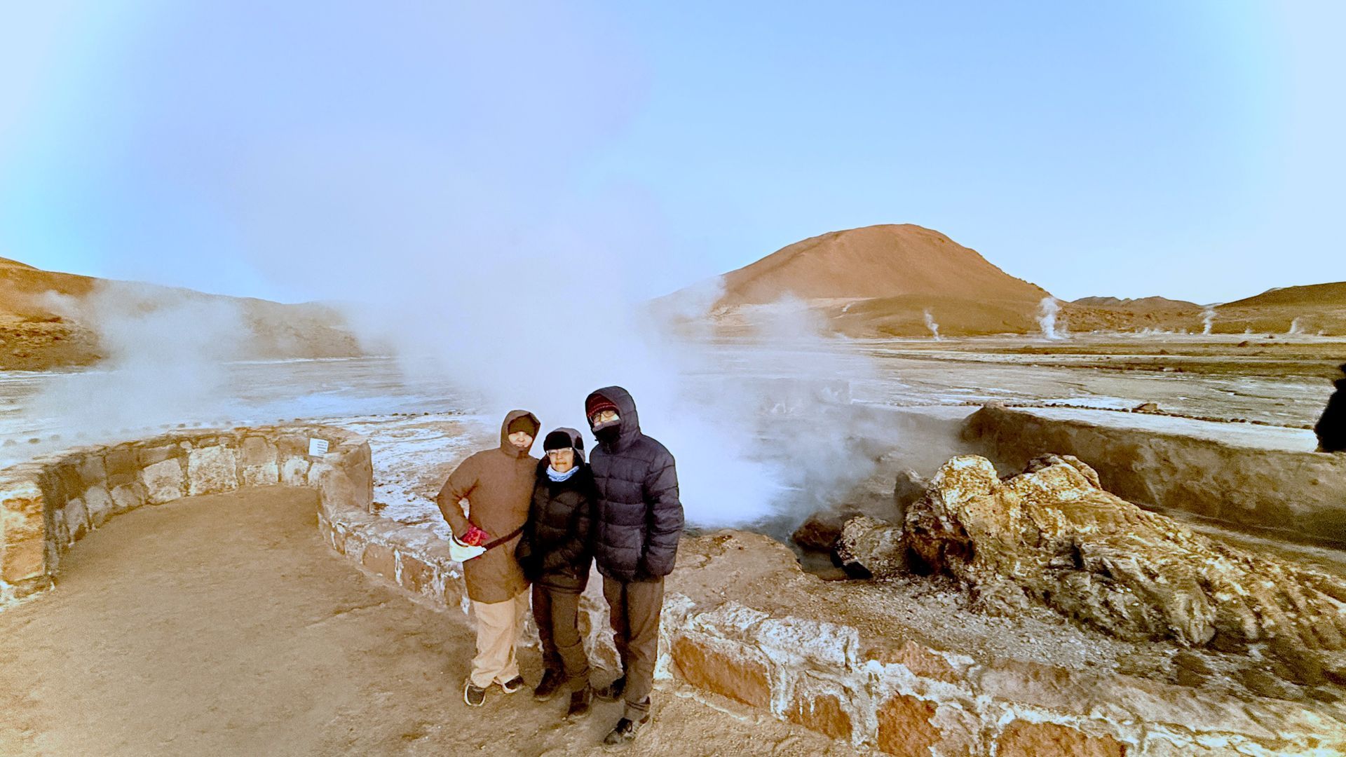Dos excursionistas bien abrigados se encuentran cerca de fumarolas geotérmicas humeantes en un paisaje rocoso y nevado.