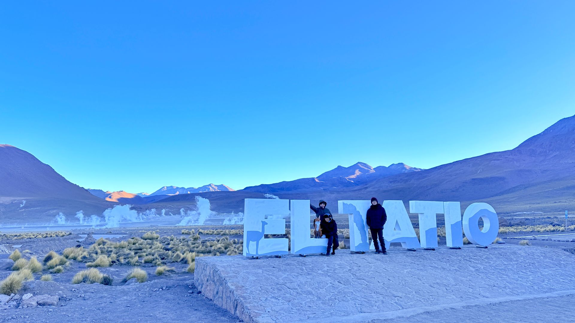 Personas posando junto a grandes letras blancas en un paisaje desértico con montañas y cielo azul.
