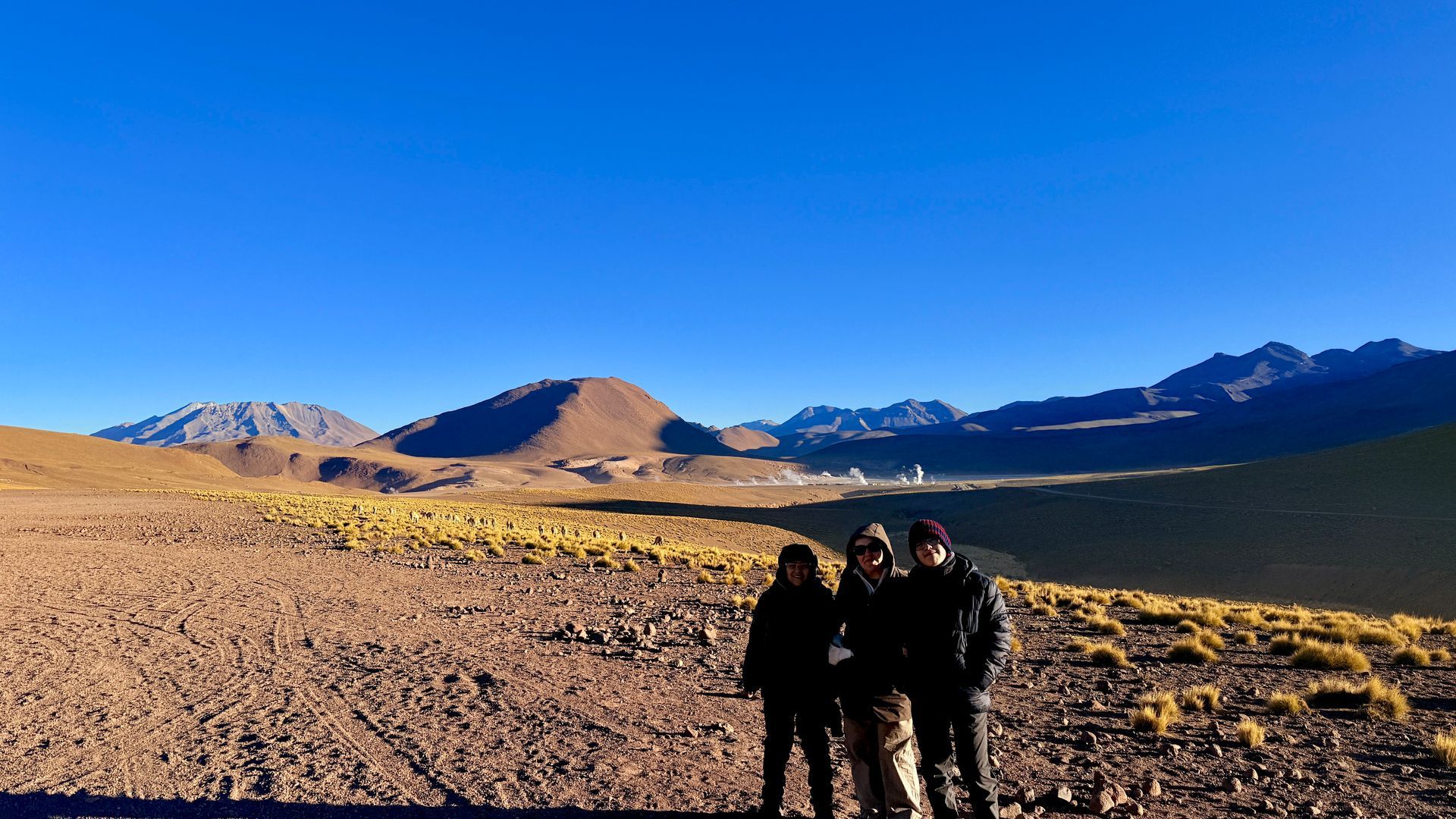 Tres personas de pie en una llanura rocosa con montañas y un cielo azul al fondo.