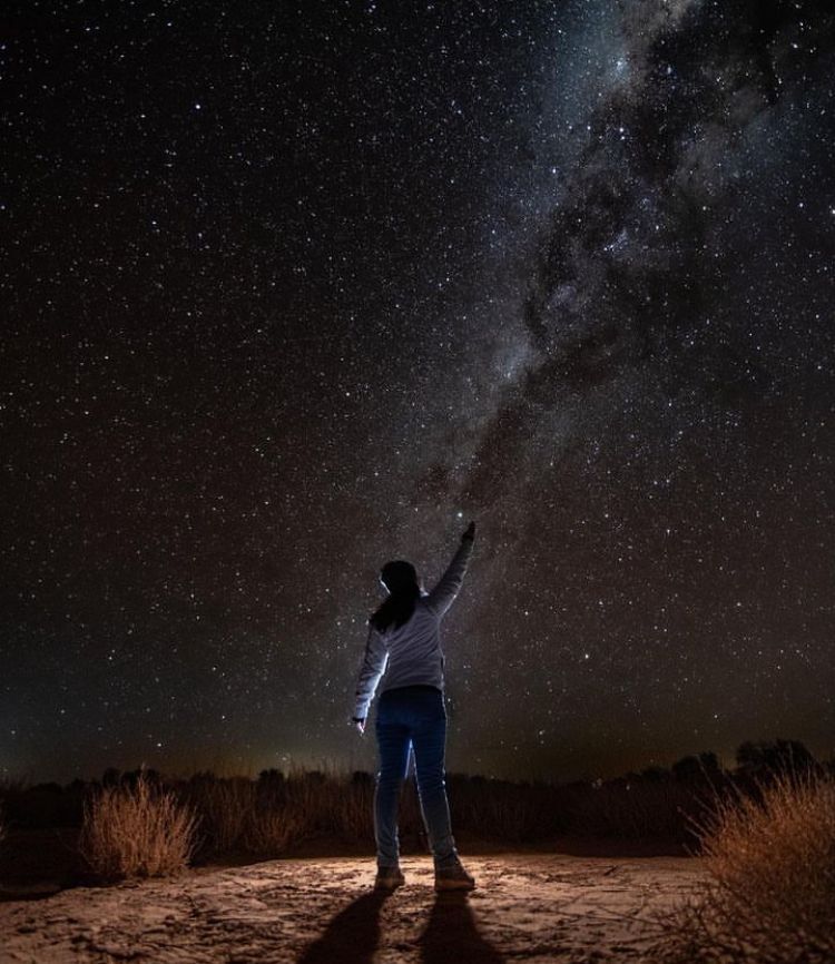 Una persona de pie bajo un cielo nocturno estrellado, señalando la Vía Láctea sobre un paisaje oscuro.