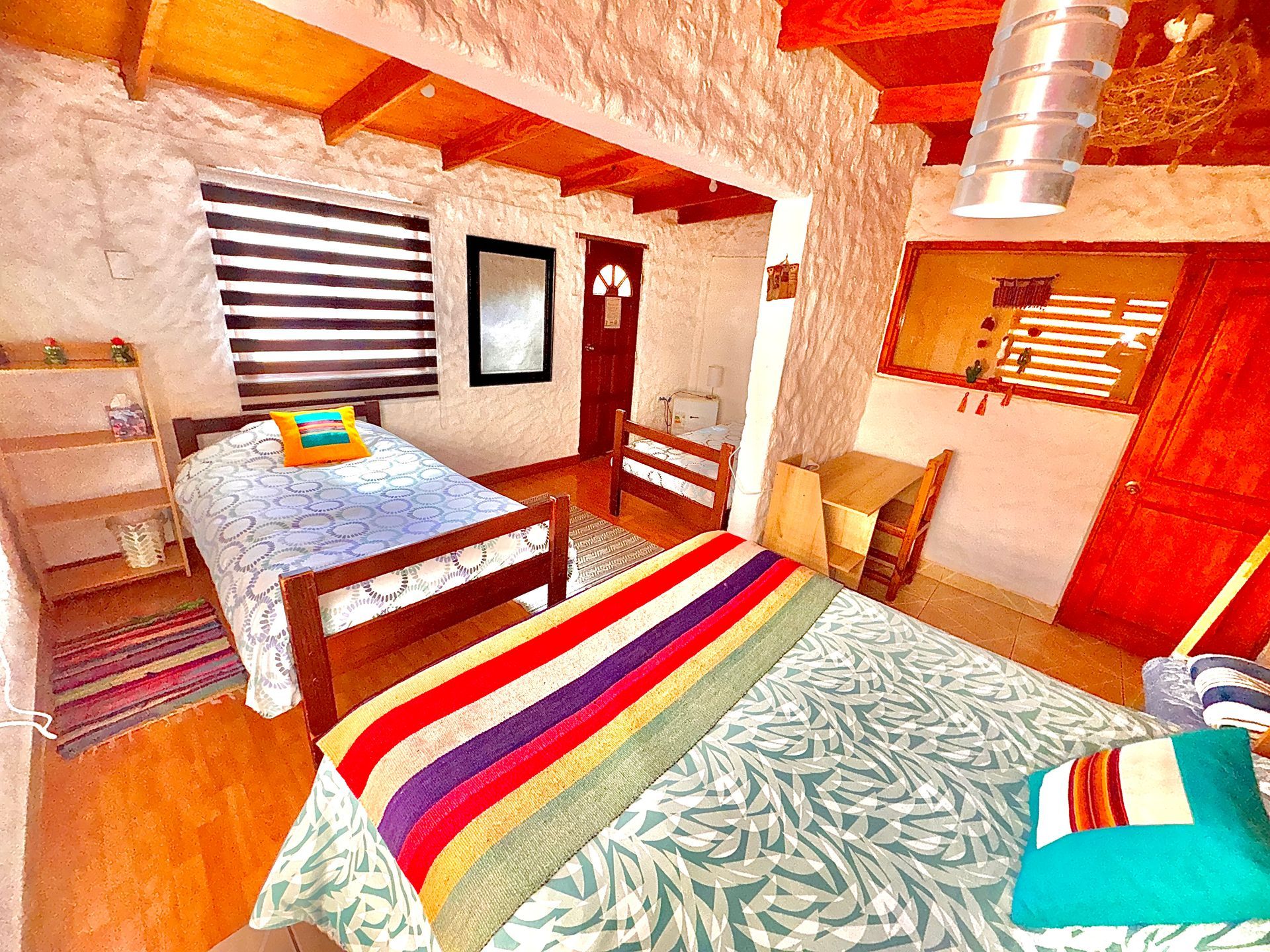 Habitación luminosa con dos camas, una manta de rayas de colores, muebles de madera y una ventana con persianas de rayas.