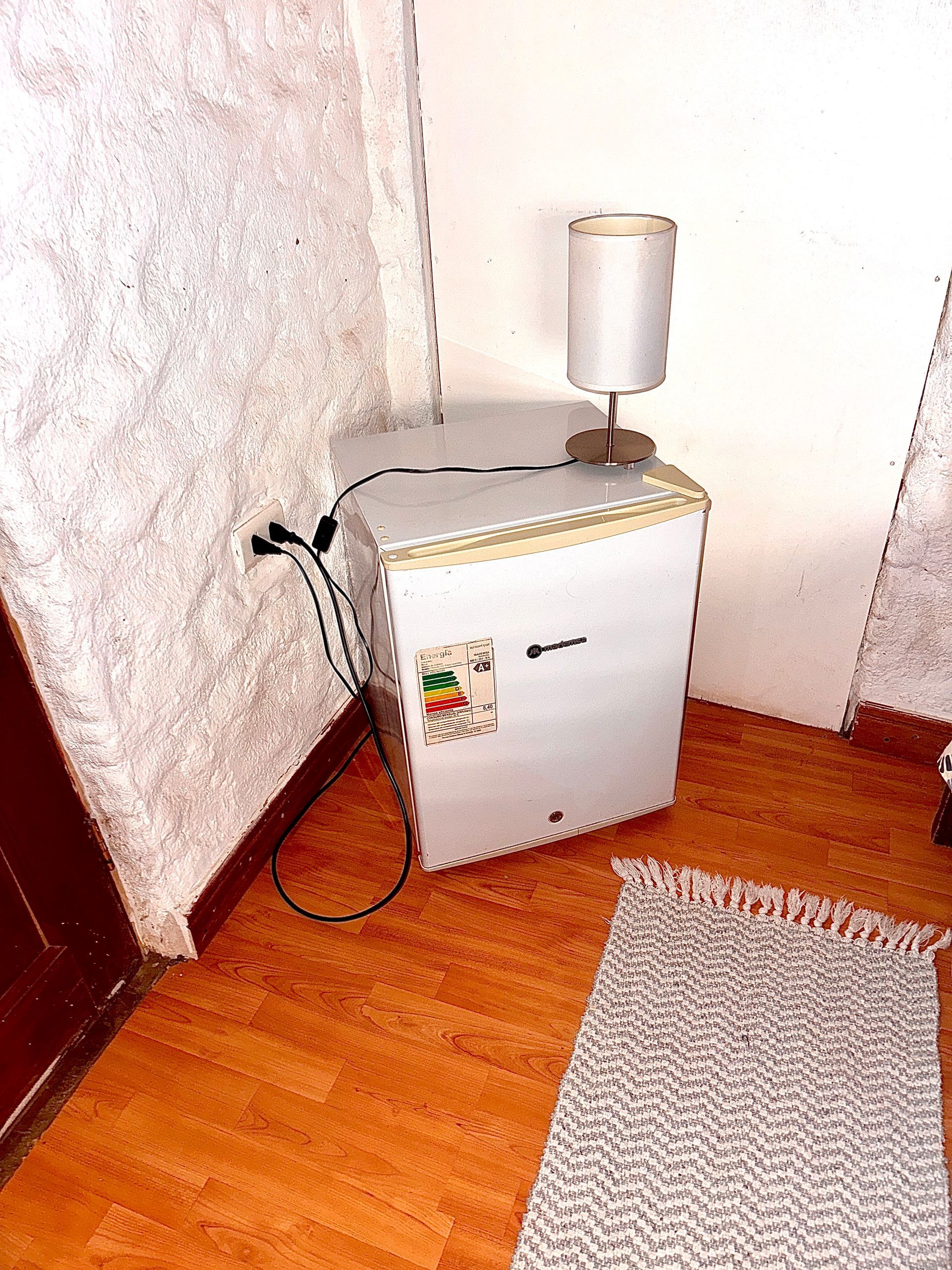Mininevera blanca pequeña junto a un enchufe de pared, con una lámpara encima y una alfombra en el suelo de madera.