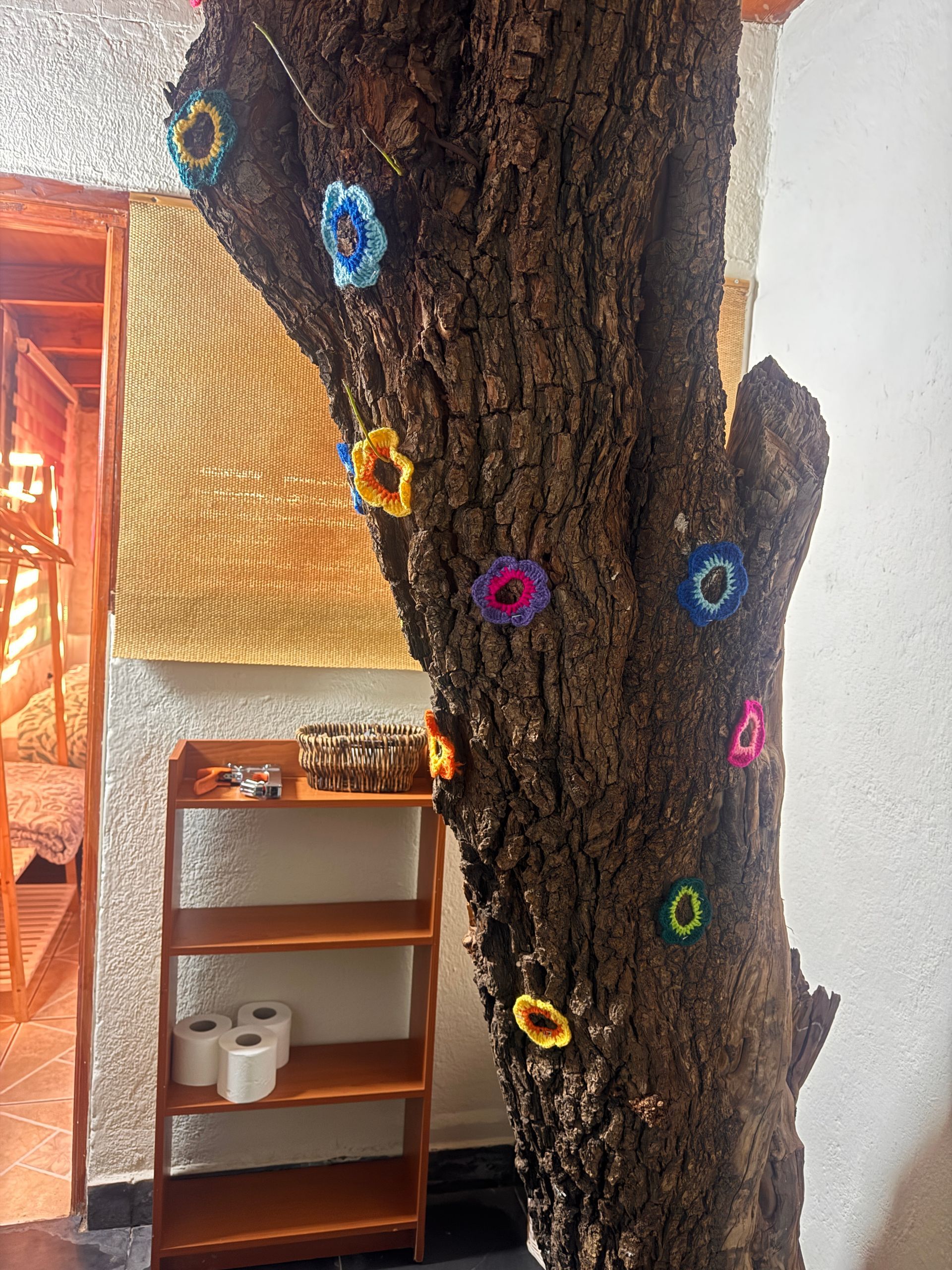 Tronco de árbol de interior decorado con coloridos adornos florales junto a un estante de madera y una pared blanca.