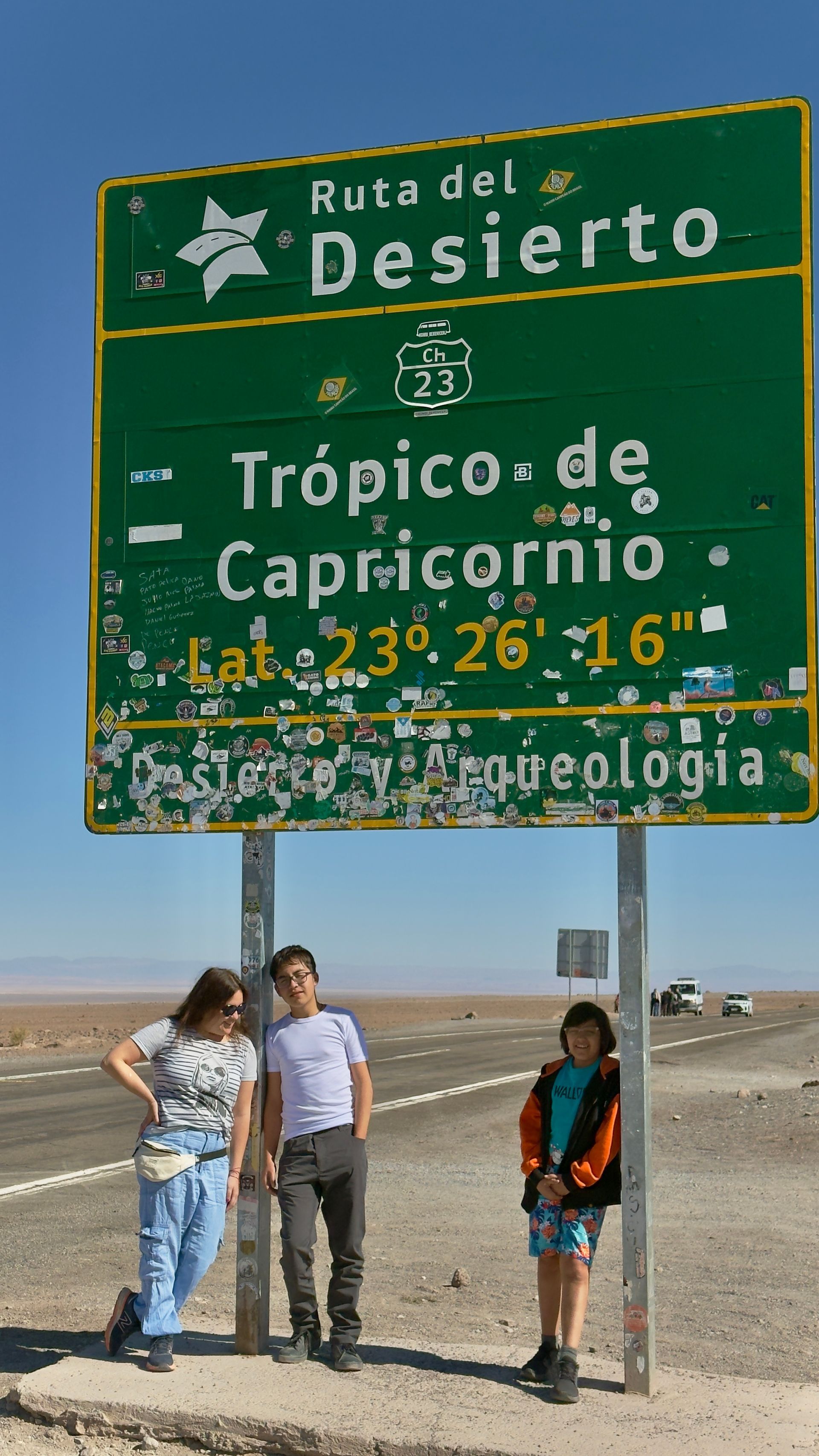 Tres personas se encuentran junto a un cartel verde que señala el Trópico de Capricornio en el desierto.