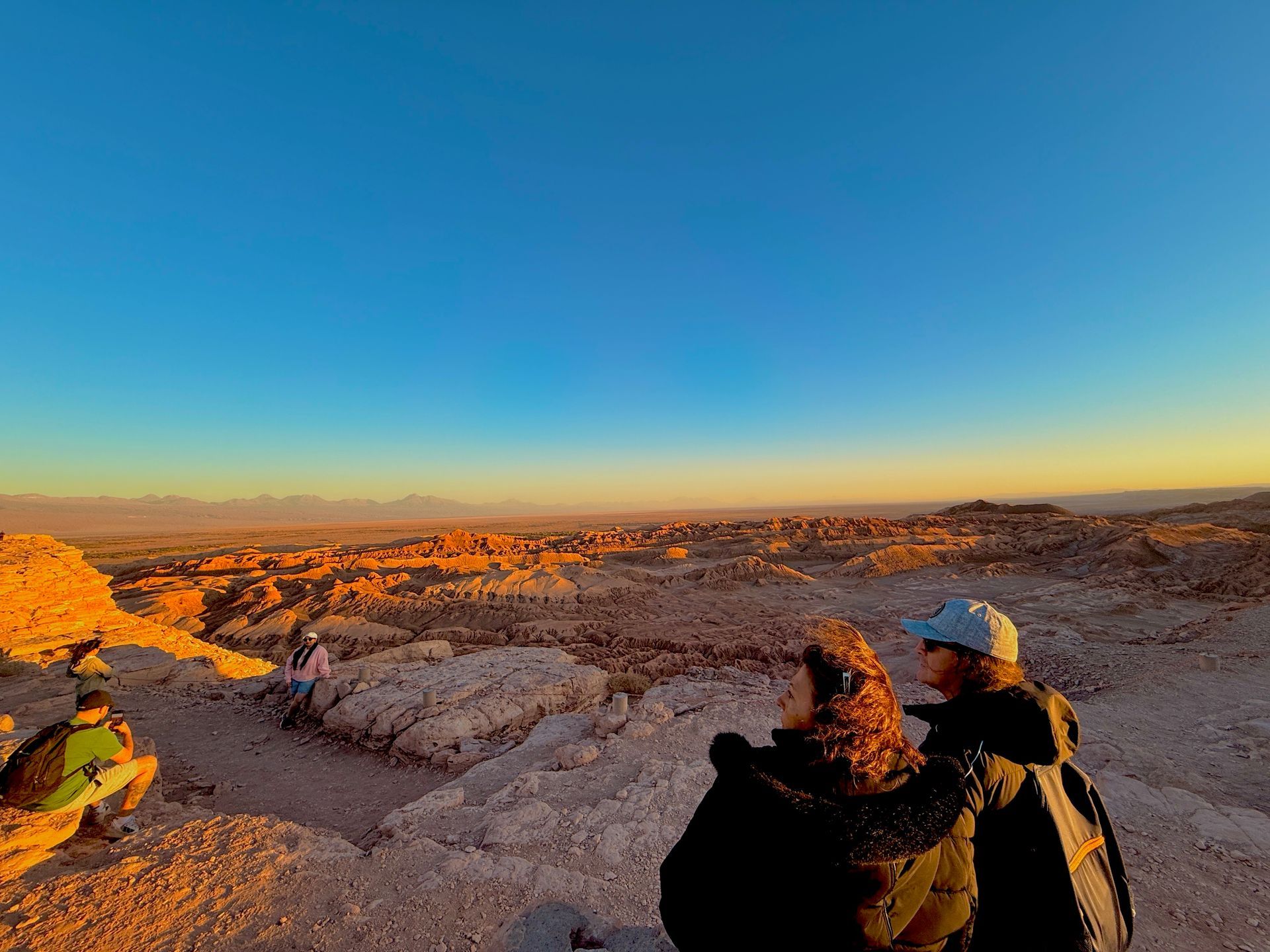 Tres excursionistas contemplan un paisaje desértico rocoso iluminado por el sol bajo un cielo azul claro.