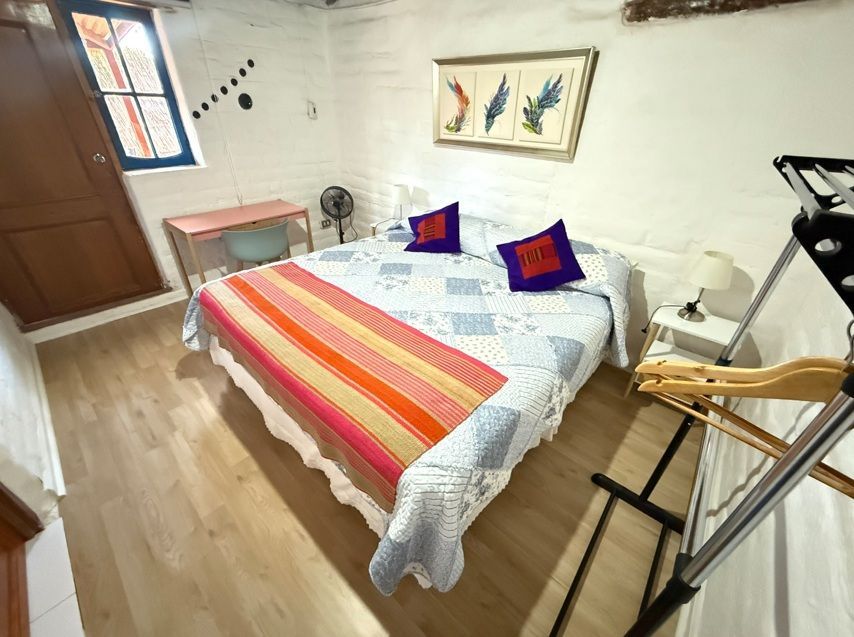 Habitación luminosa con cama doble, manta de rayas de colores y cuadros en la pared junto a la ventana y la puerta.