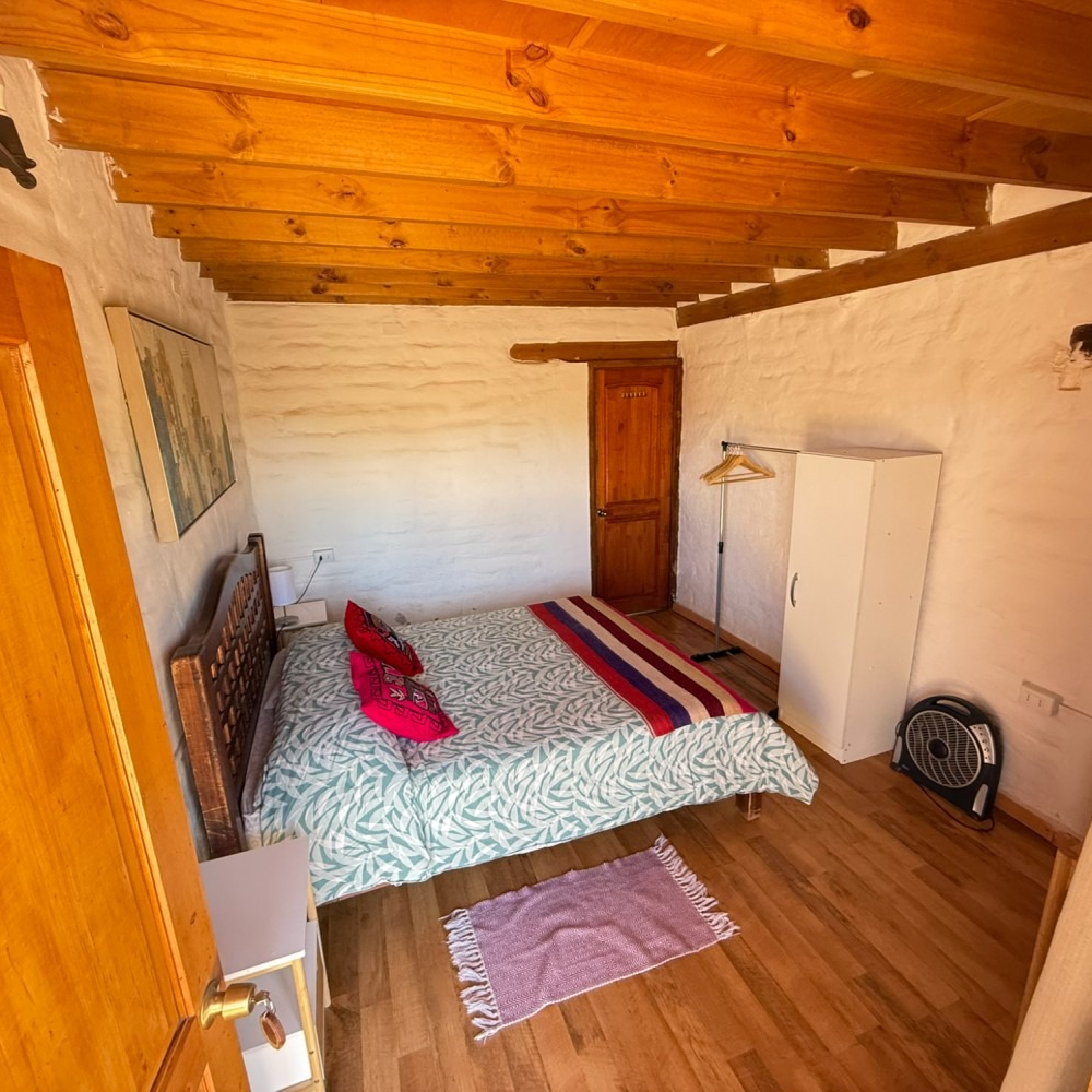 Pequeña habitación rústica con cama, techo de madera, cómoda y ventilador de pie.