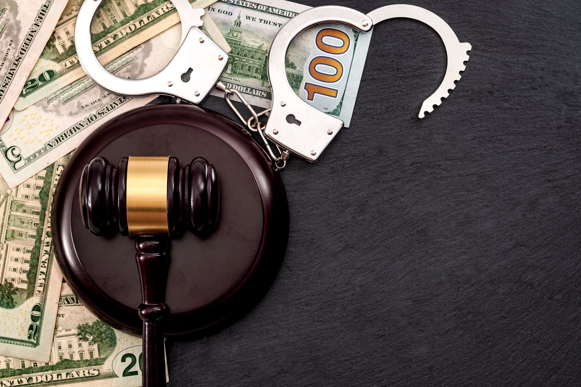 A judge’s gavel and open handcuffs resting on scattered US dollar bills against a dark surface.