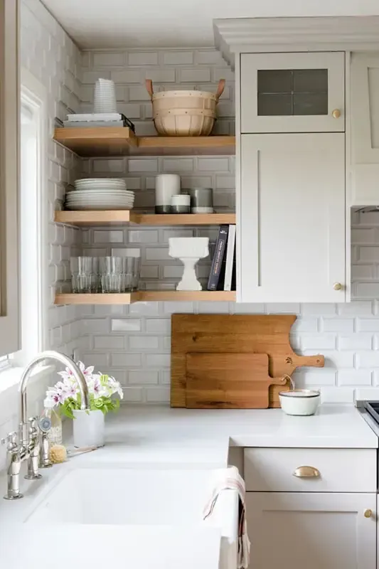 A kitchen with open shelves, white subway tile, and a farmhouse sink. Wooden cutting boards hang on the countertop.