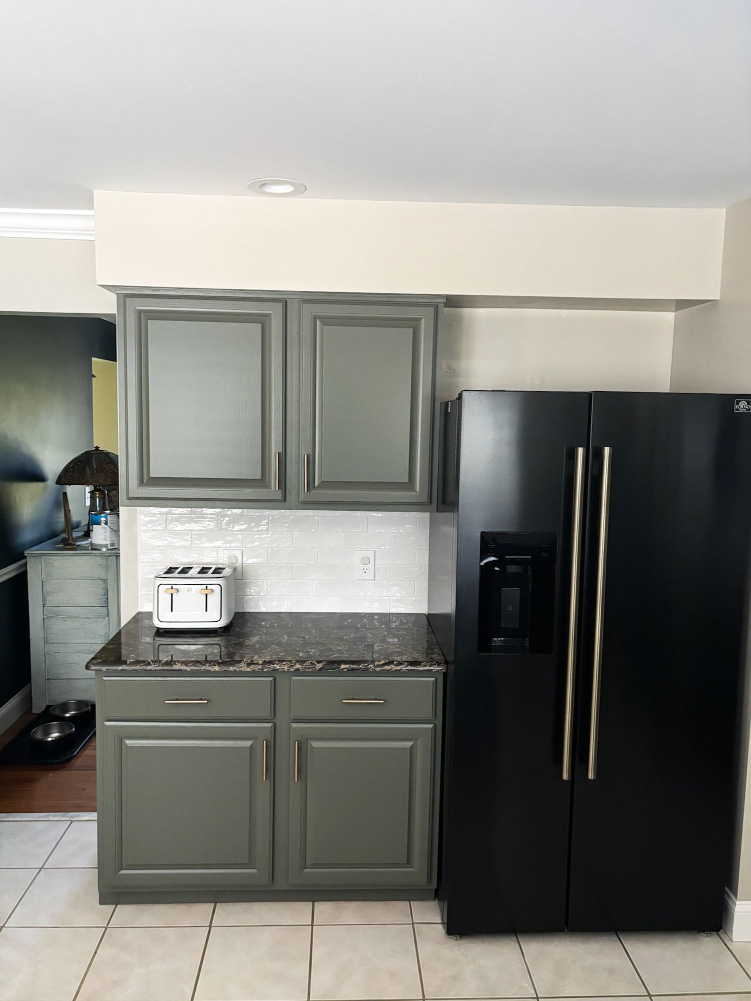 Gray kitchen cabinets and black fridge, tiled backsplash, granite countertop, toaster on counter.