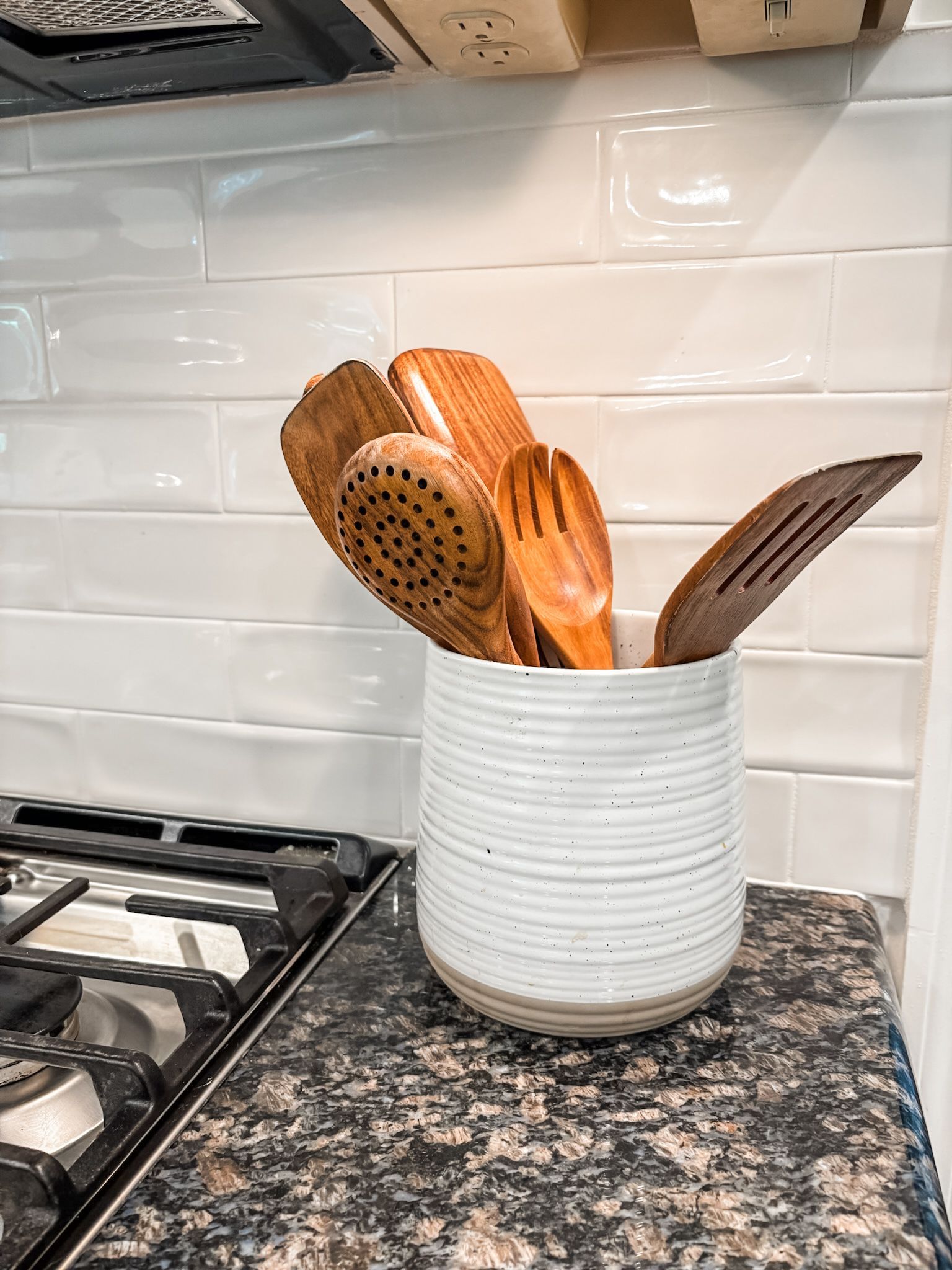 Wooden kitchen utensils in a white ceramic canister on a countertop next to a stovetop.