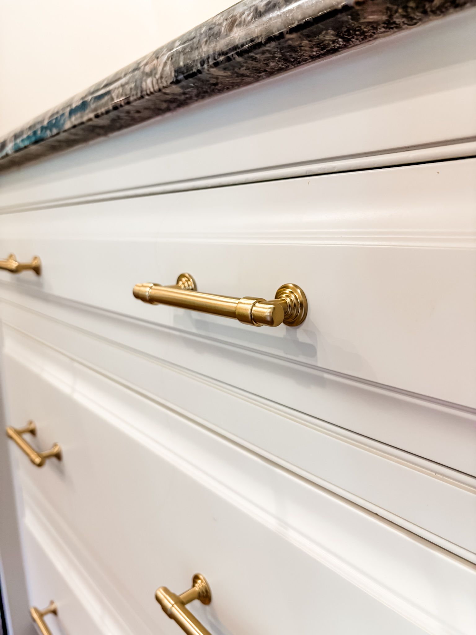 White cabinet drawers with gold handles, angled view. Dark countertop.