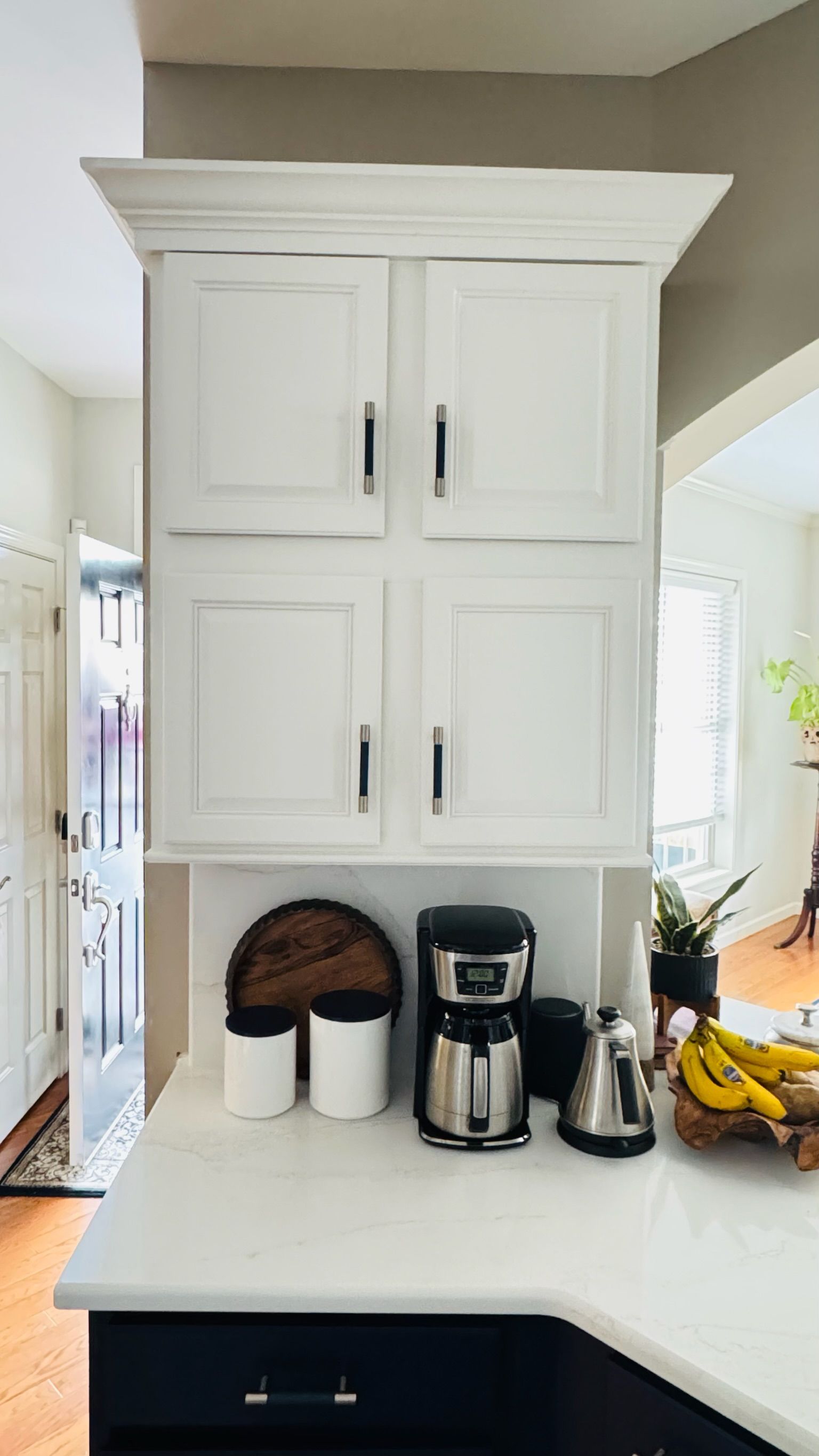 White kitchen cabinets with black hardware above a countertop with coffee maker, and bananas.
