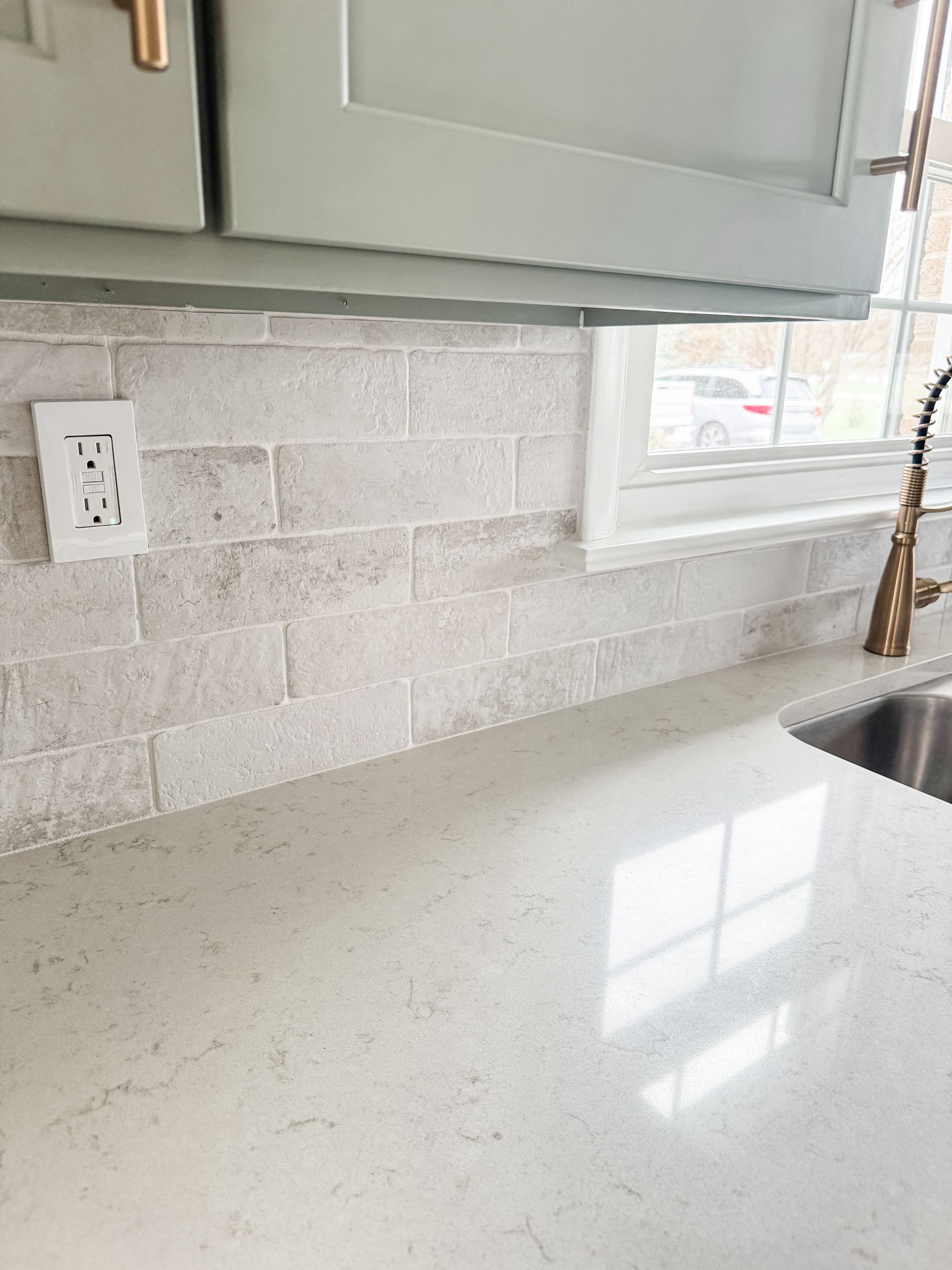 Kitchen with pale green cabinets, light gray brick backsplash, and white countertop, window.