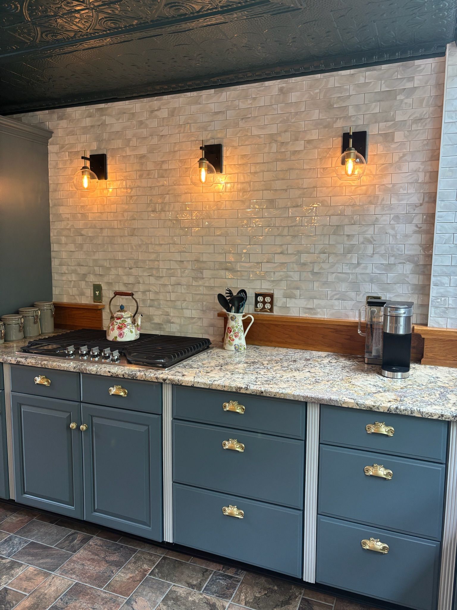 Kitchen with gray cabinets, granite countertops, brick backsplash, and three sconce lights.
