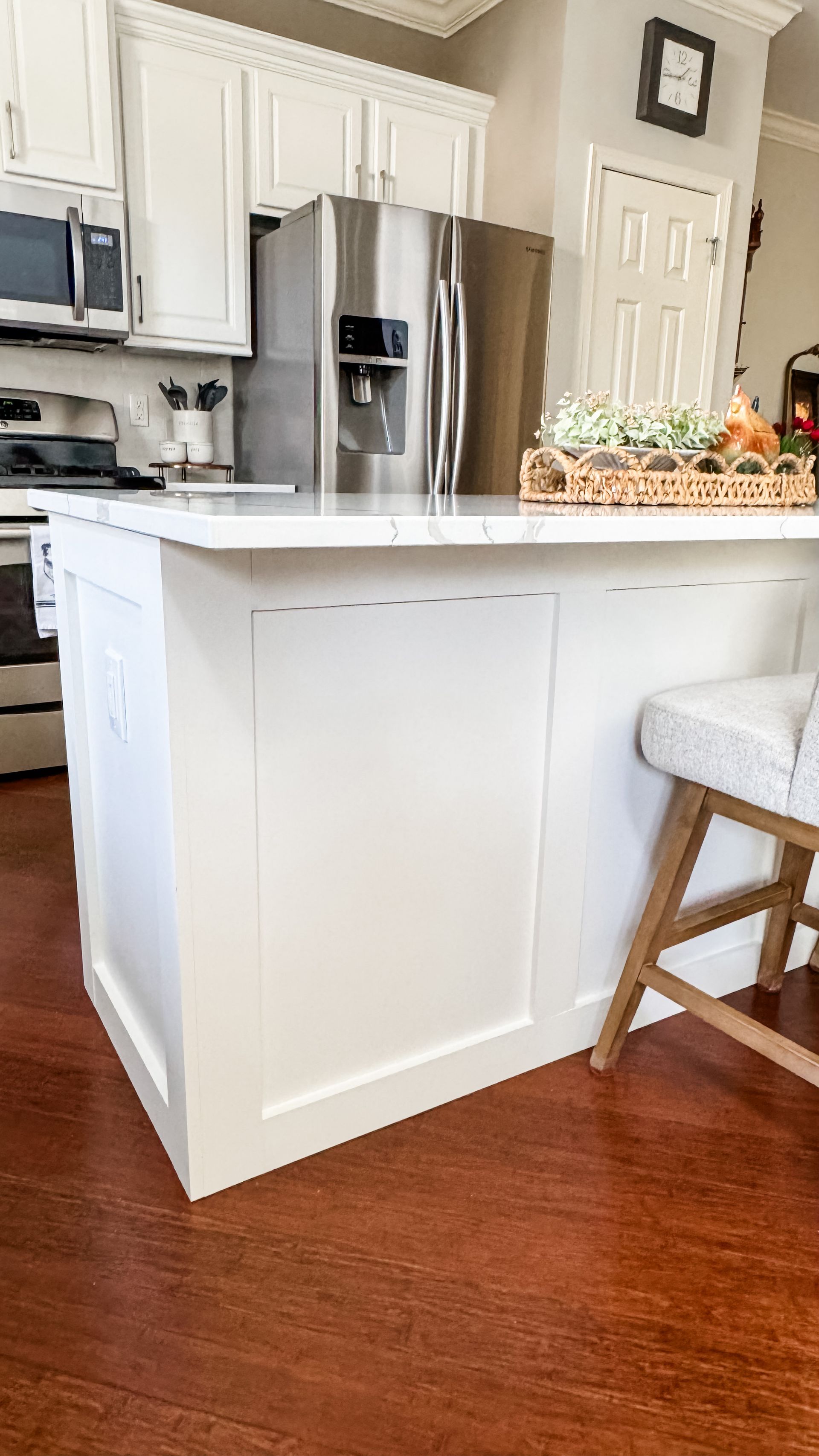 White kitchen island with marble top, stainless steel fridge, and wooden stool, on a brown floor.