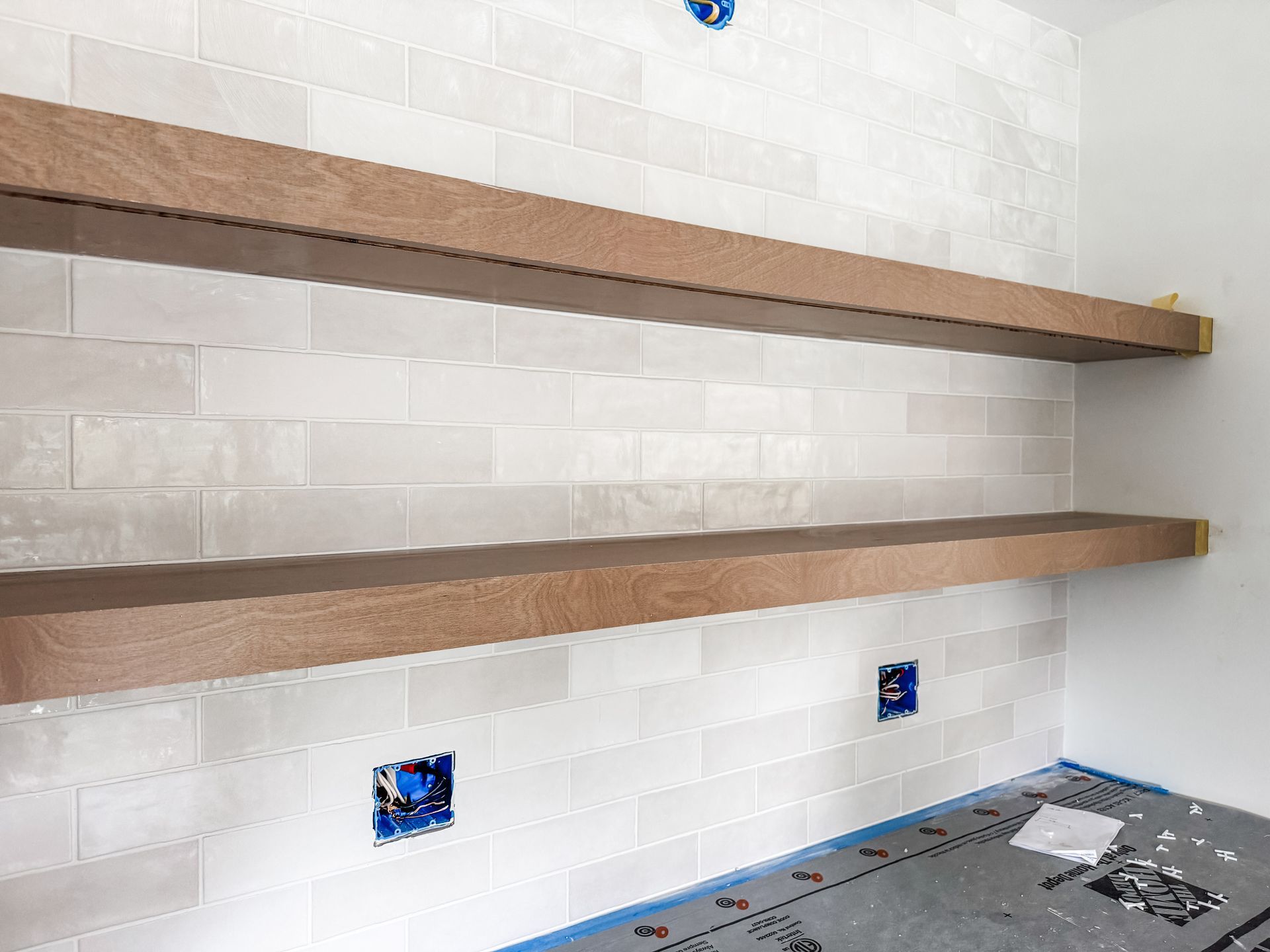 Two wooden shelves mounted on a light-tiled wall with electrical boxes.