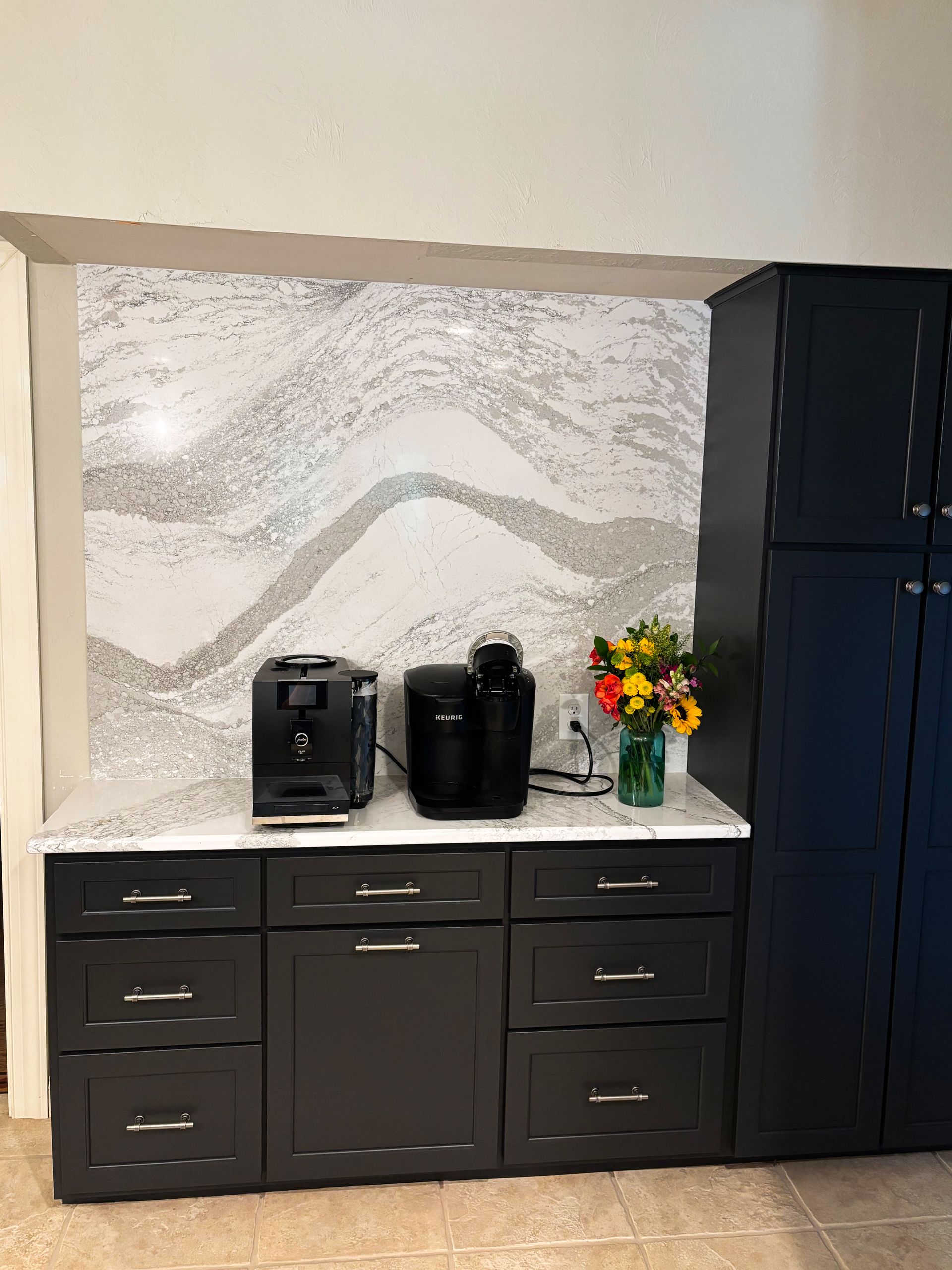 Coffee bar with black cabinets, white countertop, and marble backsplash; appliances and a vase of flowers.