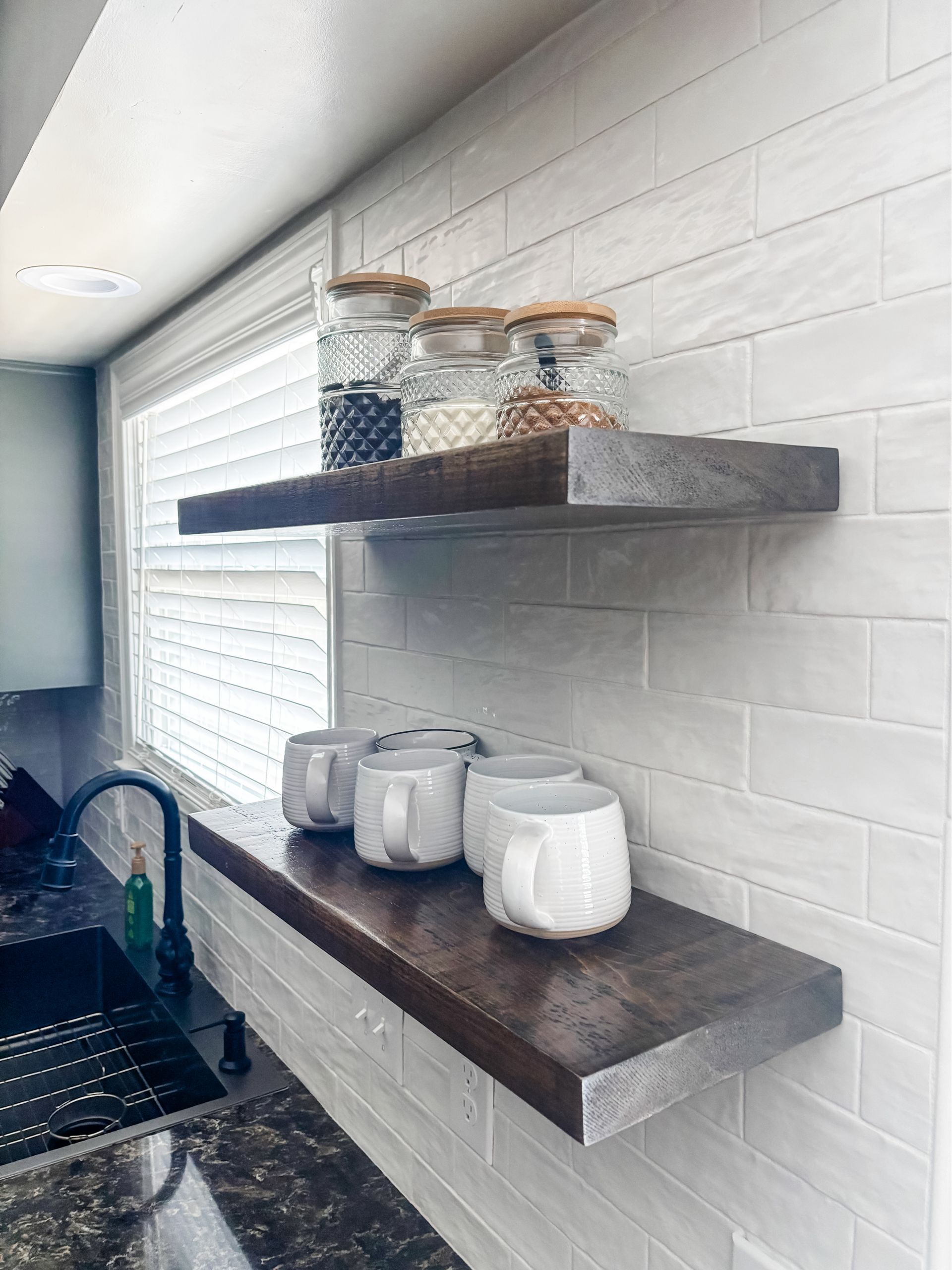 Wooden shelves in a kitchen, holding jars and mugs, against a white tiled wall.