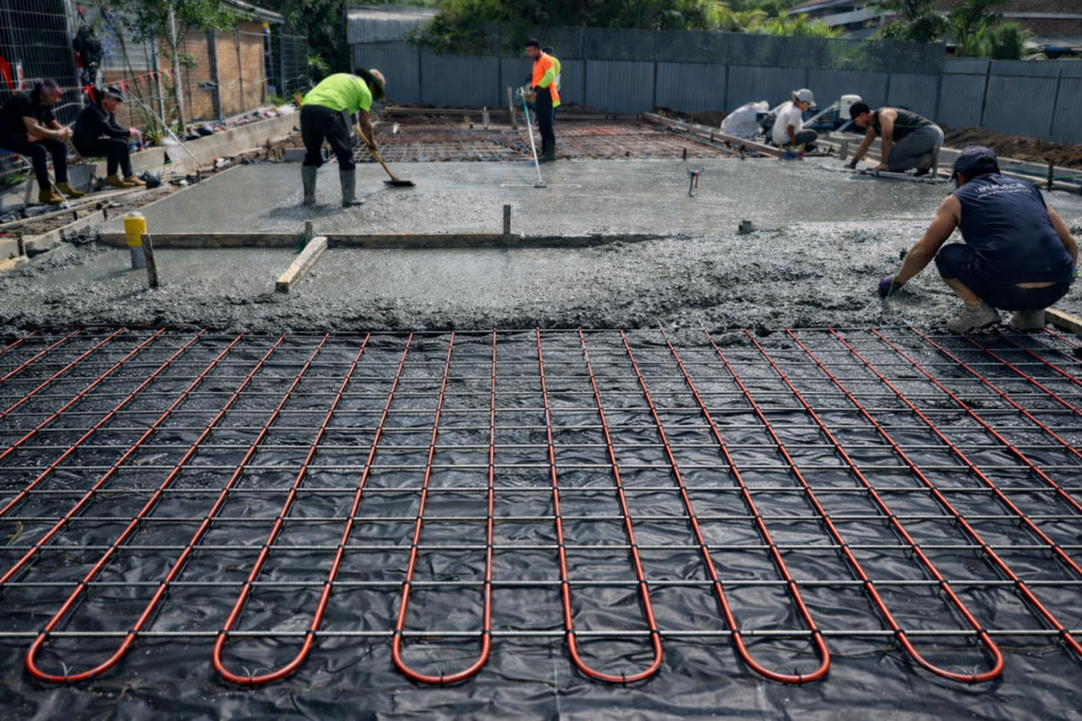 Construction workers smoothing freshly poured concrete over a radiant floor heating grid on a construction site.