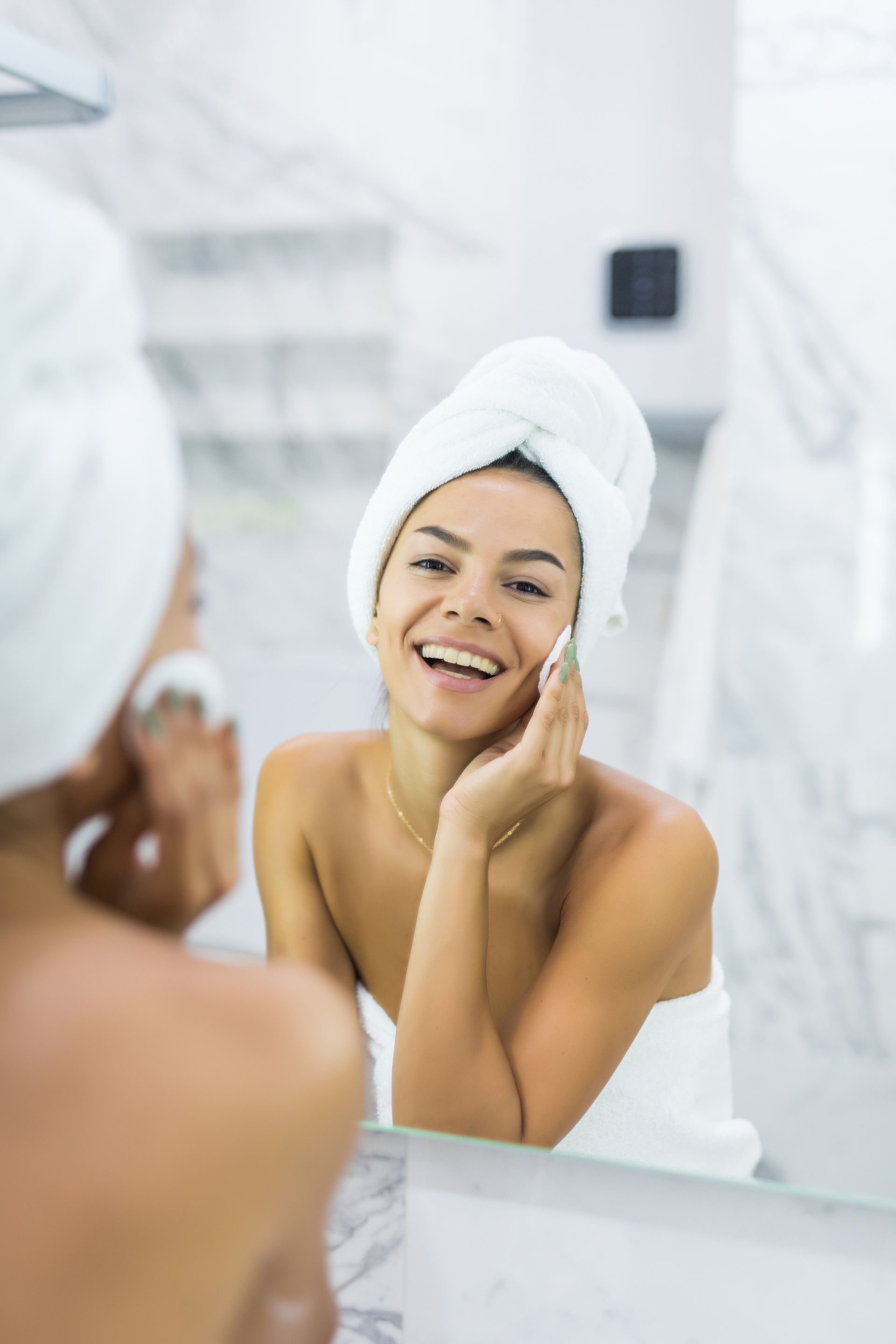 Woman in a towel smiles while cleansing her face with a cotton pad in front of a mirror.