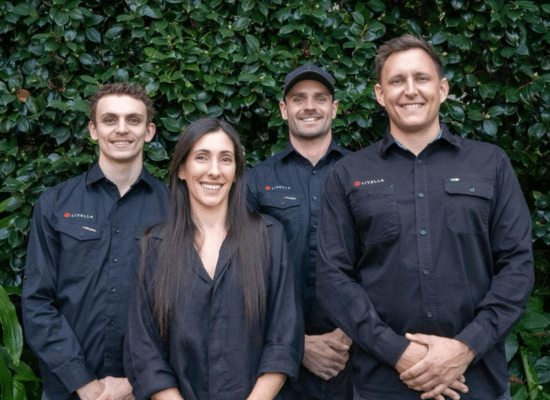 Four people in black shirts standing in front of green foliage; two men and two women are smiling.