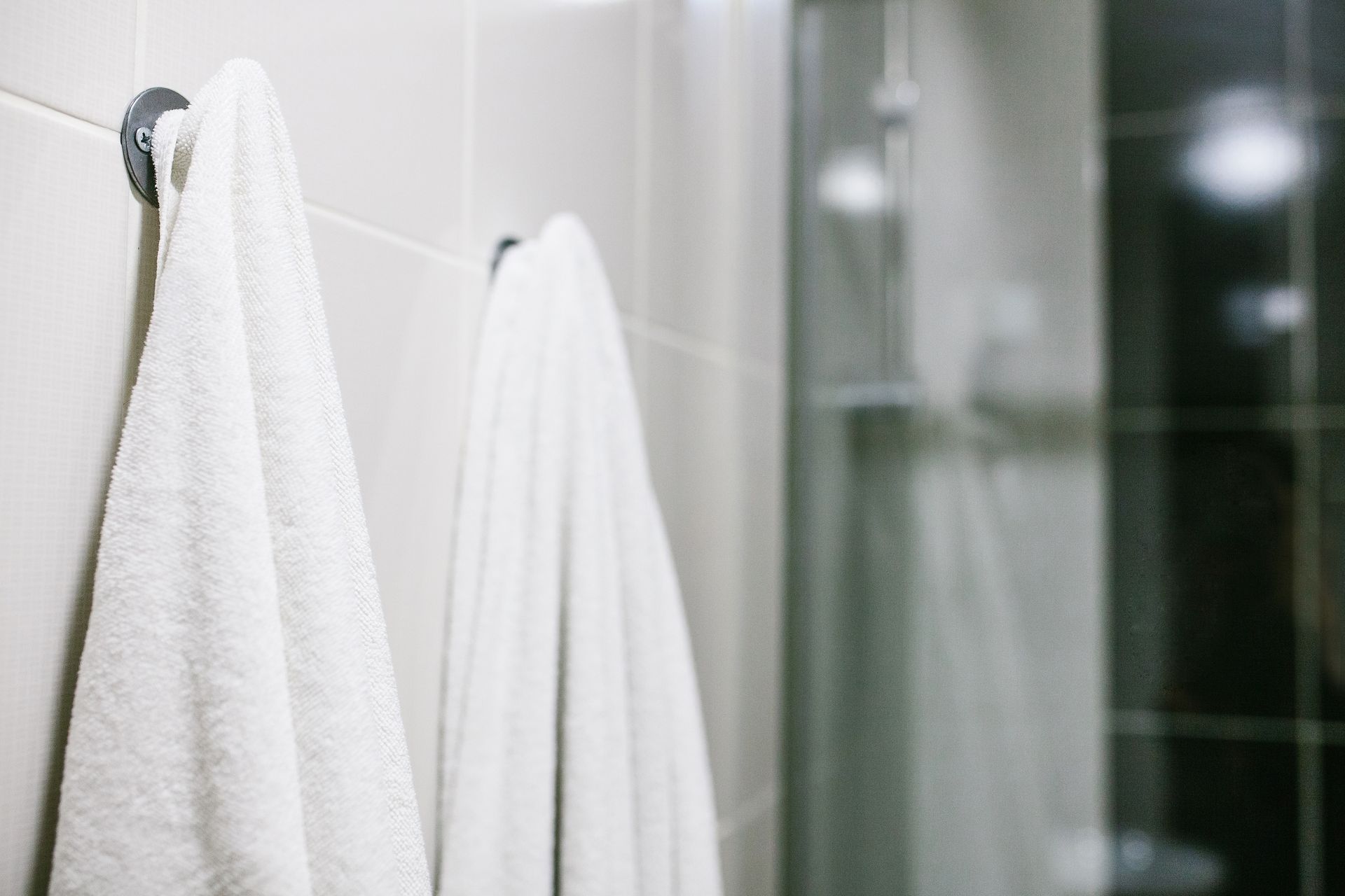 White towels hanging on silver hooks on a tiled wall, near a shower.