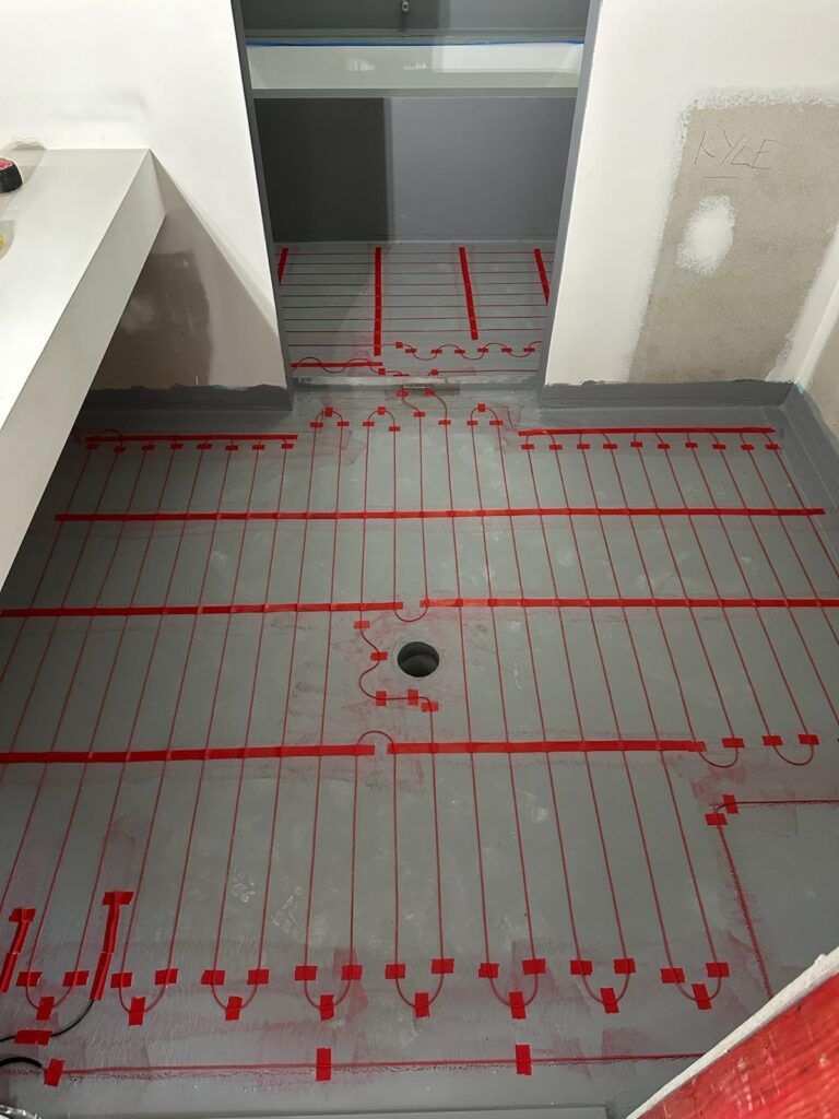 Gray bathroom floor with red heating cables, drain in center, doorway at back.
