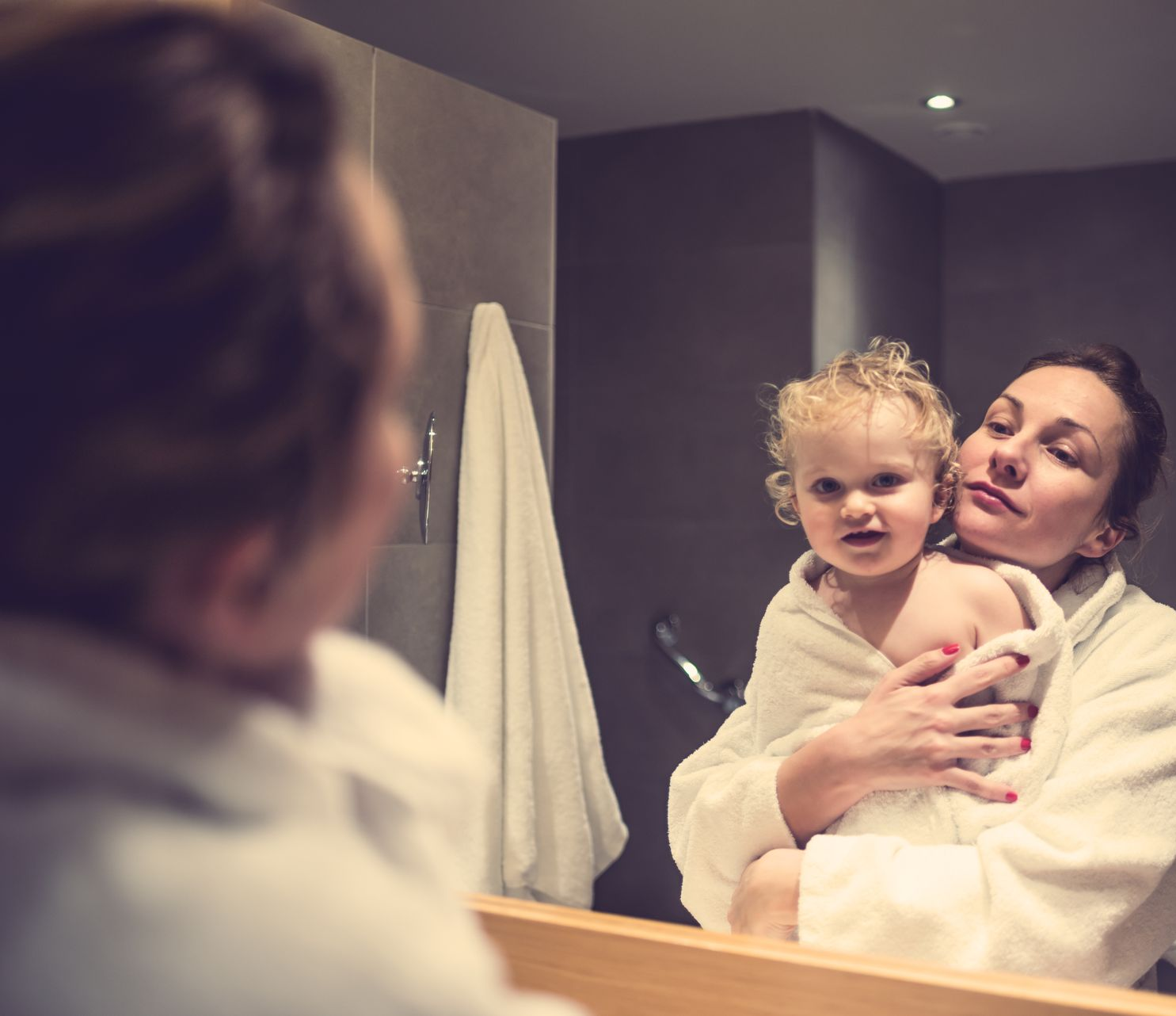Woman in a robe holds a child wrapped in a towel in front of a bathroom mirror.