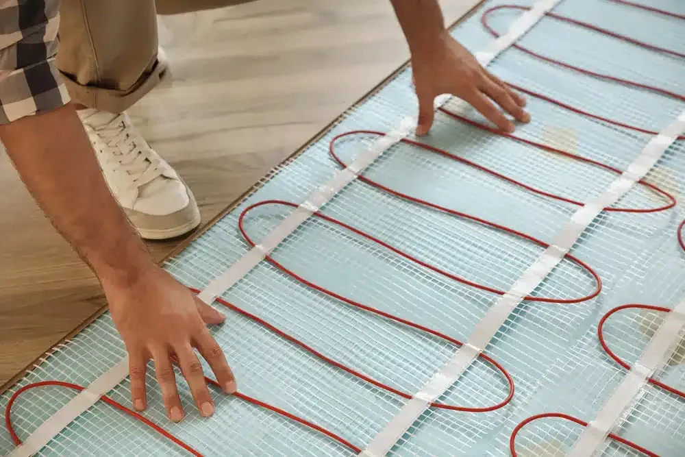Person installing radiant floor heating system, hands on a blue mat with red heating cables.