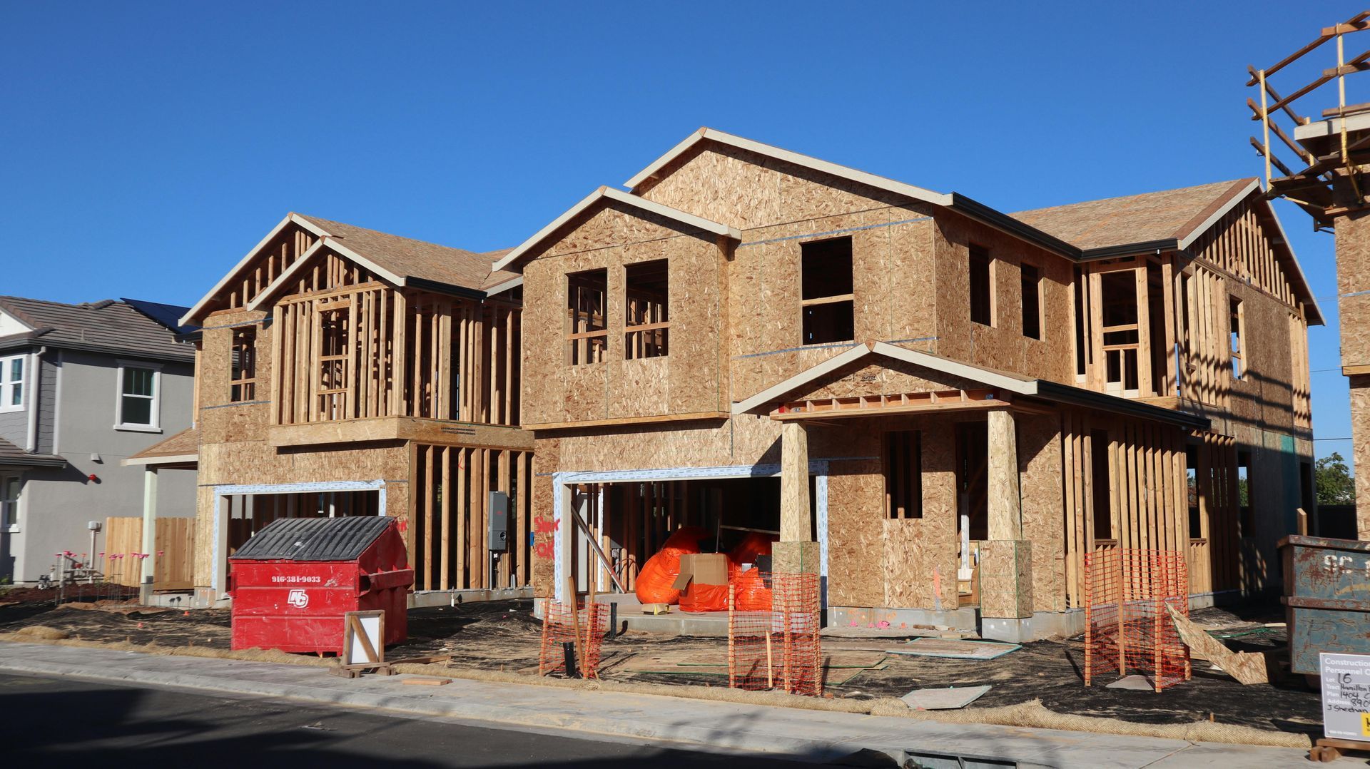 Two-story house under construction with exposed wood framing, red dumpster, and orange barriers under a blue sky
