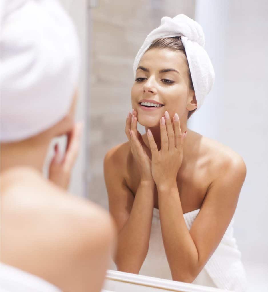 Woman in towel looking at herself in the mirror, touching her face.