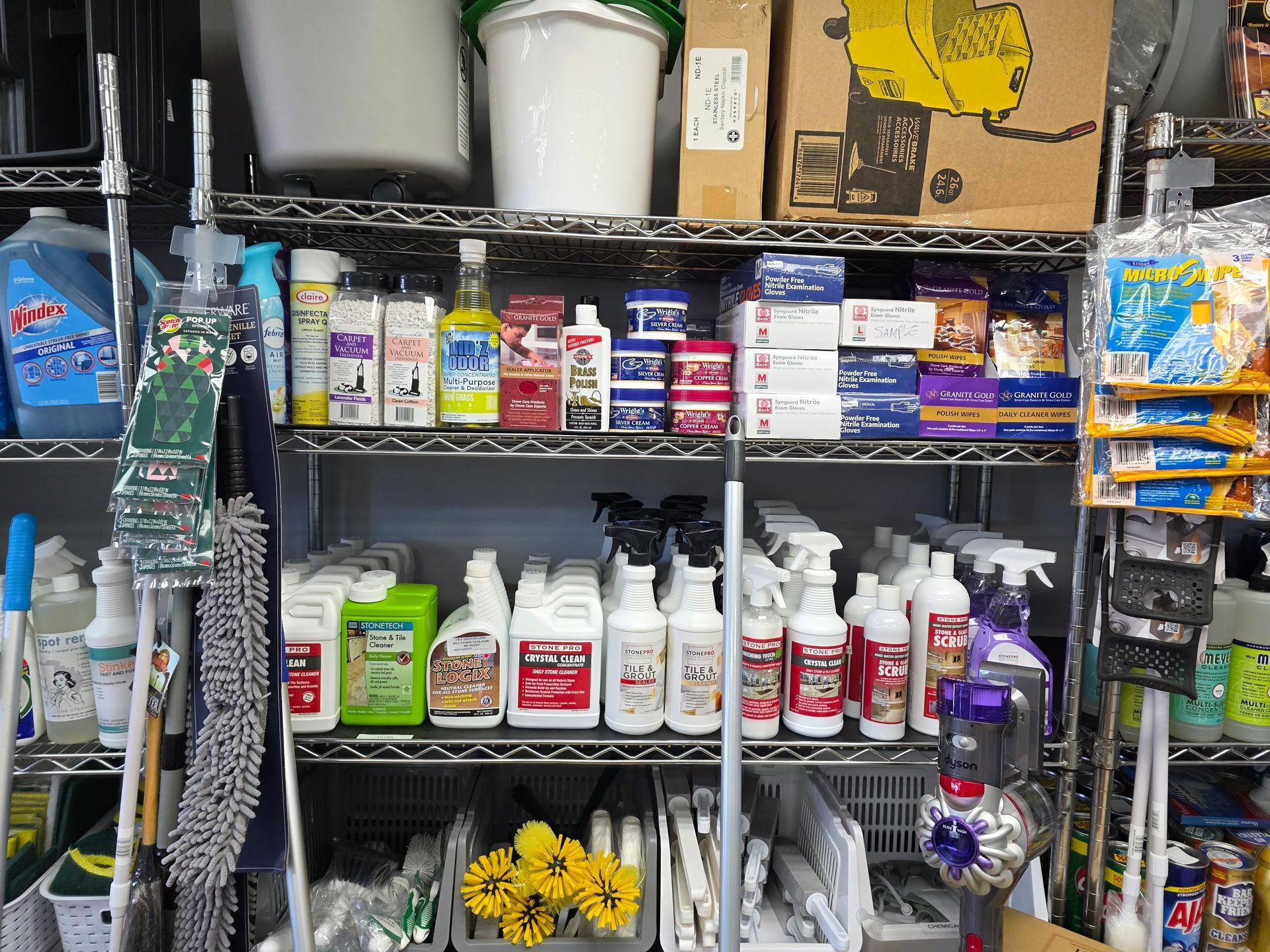 Wire shelving unit stocked with various cleaning supplies, including bottles, boxes, and brooms in a storage area.