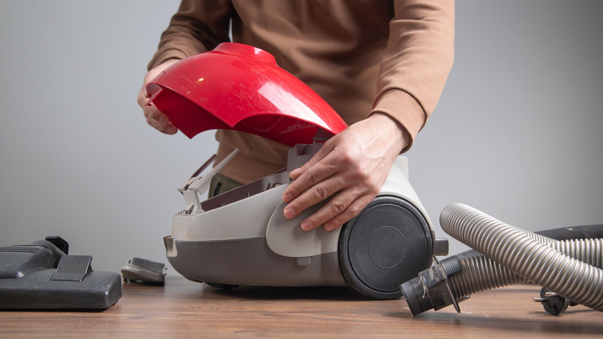 A person in a brown shirt disassembles a red and gray vacuum cleaner on a wooden table.