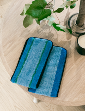 Two blue and green microfiber mop pads resting on a light wooden table next to a vase and a candle.