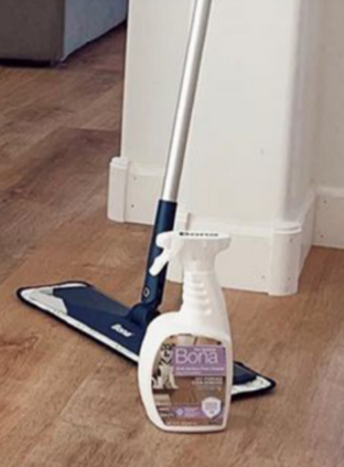A Bona floor mop and a spray bottle of Bona hardwood floor cleaner sitting on a light-colored wood floor near a wall.