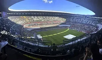 Vista interior panorámica de un gran estadio con gradas que rodean un campo verde bajo un cielo azul despejado.